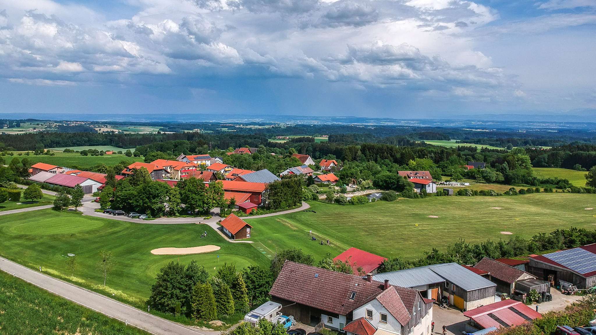 Luftbild zeigt eine hügelige Golfanlage mit Bunkern und Gebäuden, dahinter weite Landschaft mit Feldern, Wald und Alpenblick unter einem dramatischen Wolkenhimmel