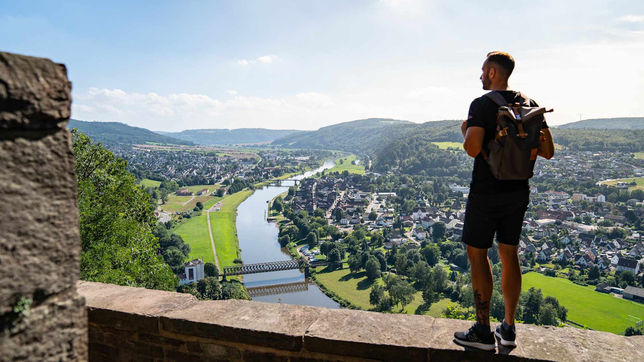 Ein Wanderer steht auf der Mauer einer Burg und schaut über die weite Landschaft, die sich unter ihm ausbreitet. Es ist Sommer, ein Fluss schlängelt sich durch die Landschaft und mehrere Ortschaften liegen am Ufer.