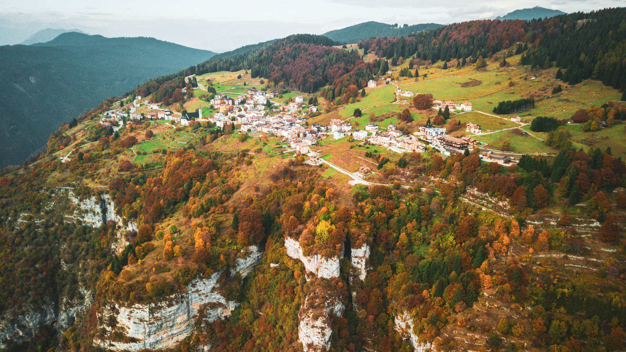 Luftaufnahme des Dorfes Luserna in Italien, das auf einem grünen Hochplateau liegt, umgeben von herbstlich gefärbten Wäldern und weißen Felswänden.