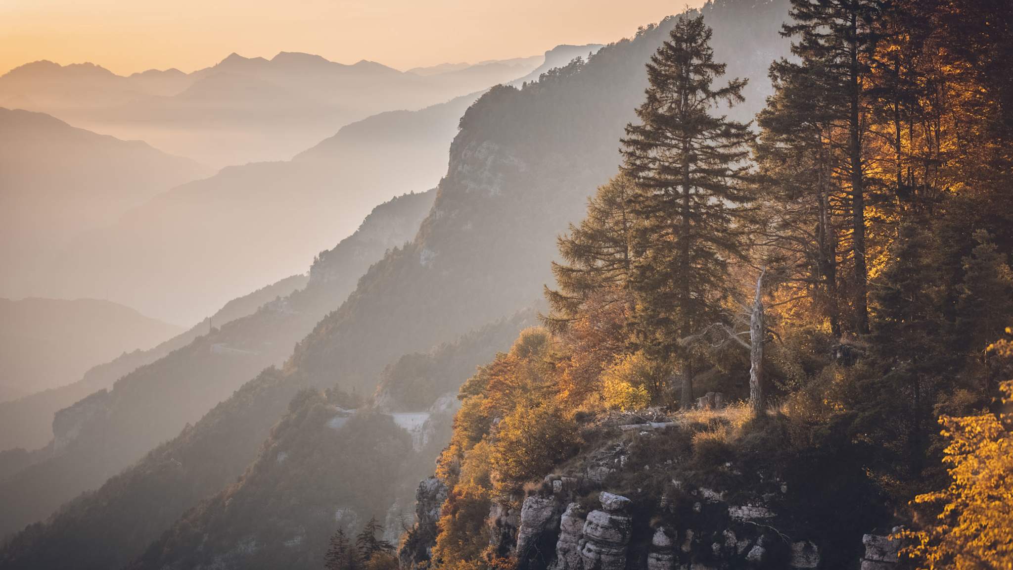 Blick auf das Vigolana-Massiv bei Sonnenuntergang: im Vordergrund herbstlich gefärbte Bäume und Felsen, im Hintergrund ziehen sich Bergketten in goldenem Licht und Dunst.