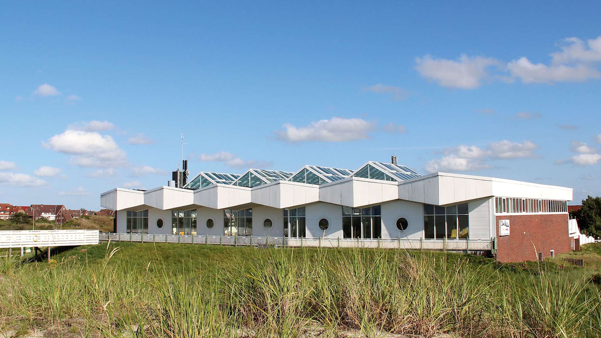 Moderne Therme mit markanter Dachstruktur und runden Fenstern, gelegen inmitten der Dünenlandschaft von Baltrum, mit Holzsteg und Blick auf die umliegende Inselarchitektur.