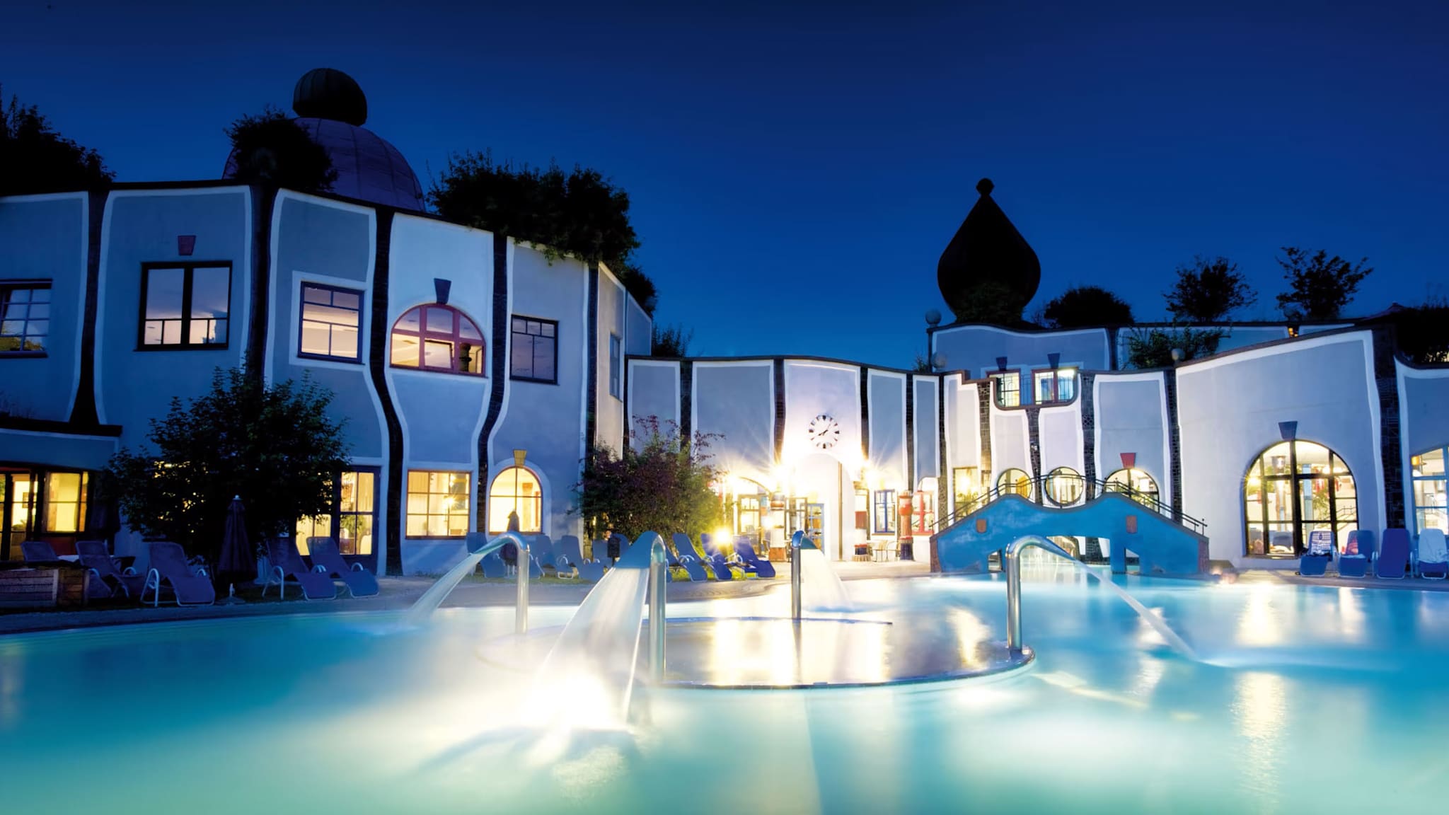 Außenpool im Rogner Bad Blumau mit warmem Licht, Wasserfontänen und geschwungener Architektur im Hintergrund.