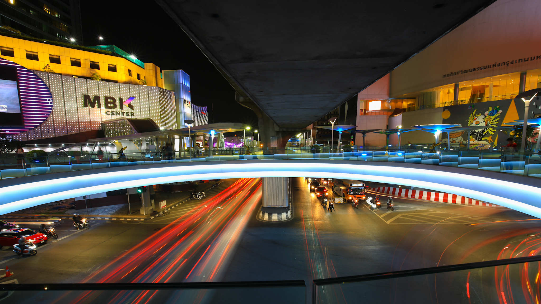Blick durch eine große, kreisrunde Bodenöffnung auf eine Straßenkreuzung in einem Tunnel unter das BTS-Skytrain-Station "Siam" und vor dem MBK Shoppingcenter.
