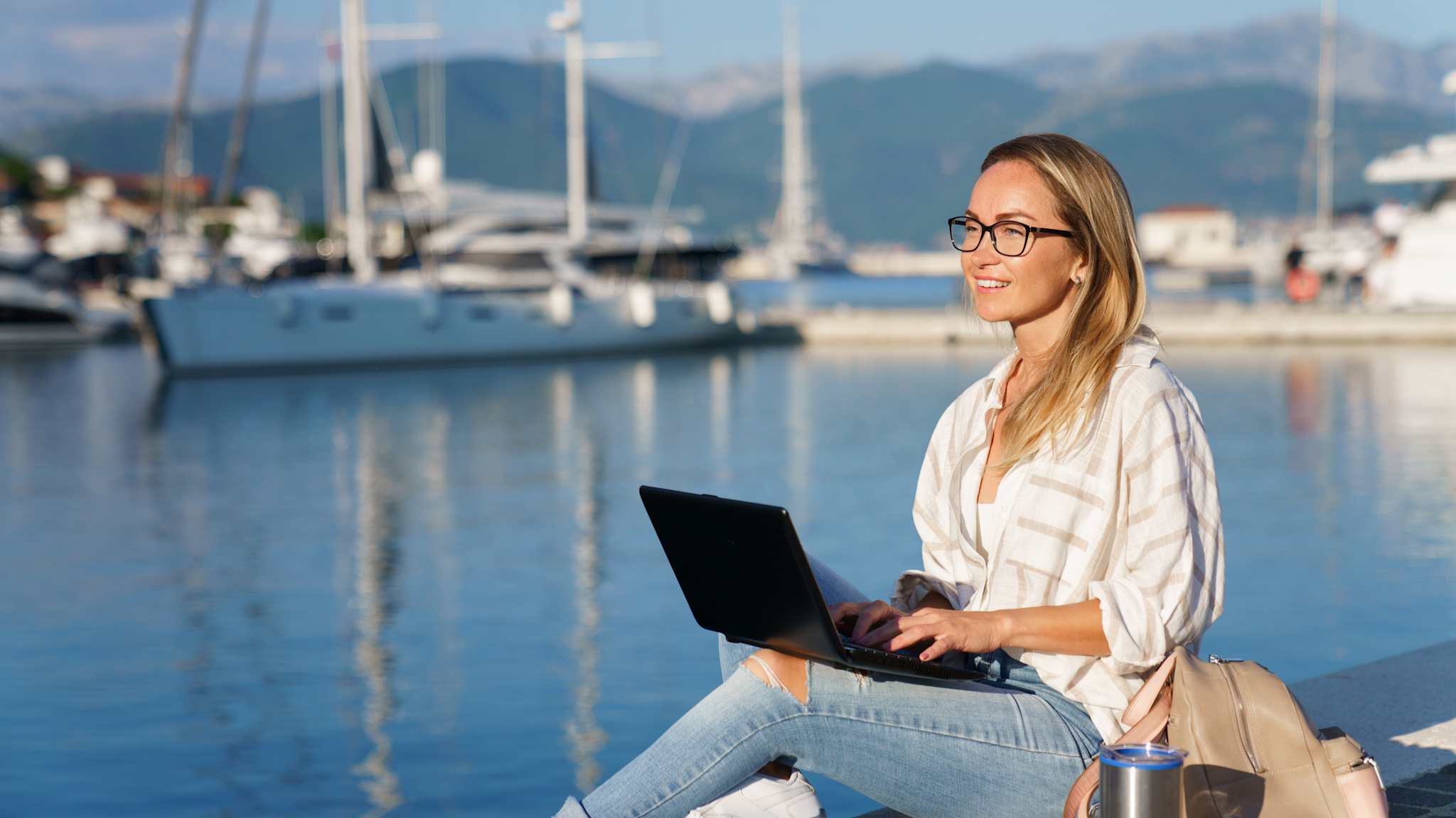 Eine Frau sitzt mit einem Laptop und Rucksack an einem Hafen am Meer mit Bergen im Hintergrund.
