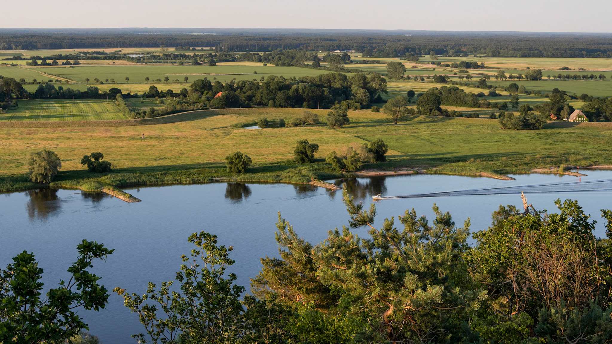 Blick über eine grüne flache Landschaft. Im Vordergrund fließt ein breiter Fluss, auf dem ein kleines Boot fährt.