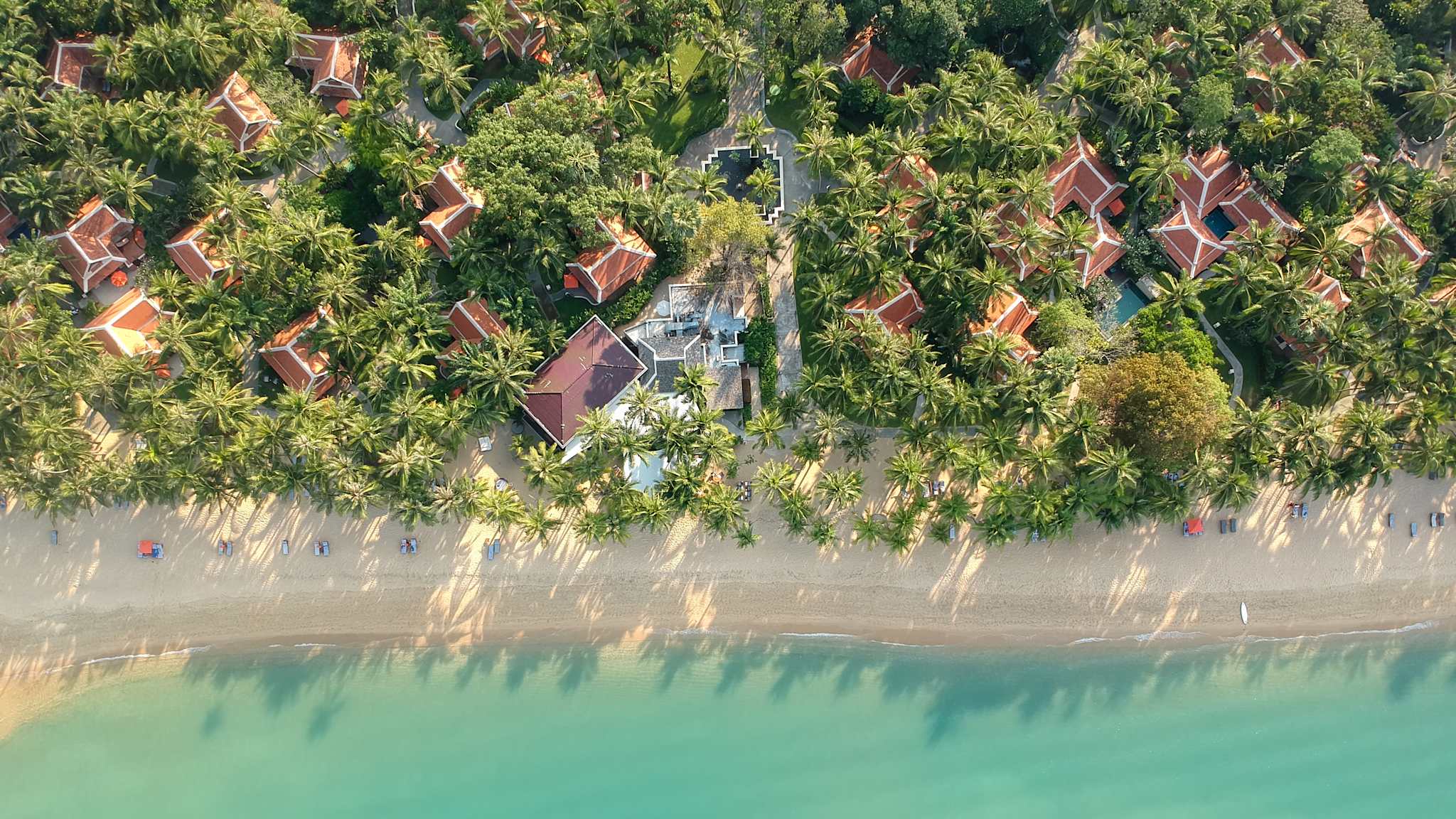 Blick von oben auf einen Strand mit Häusern und Bäumen sowie klarem Wasser