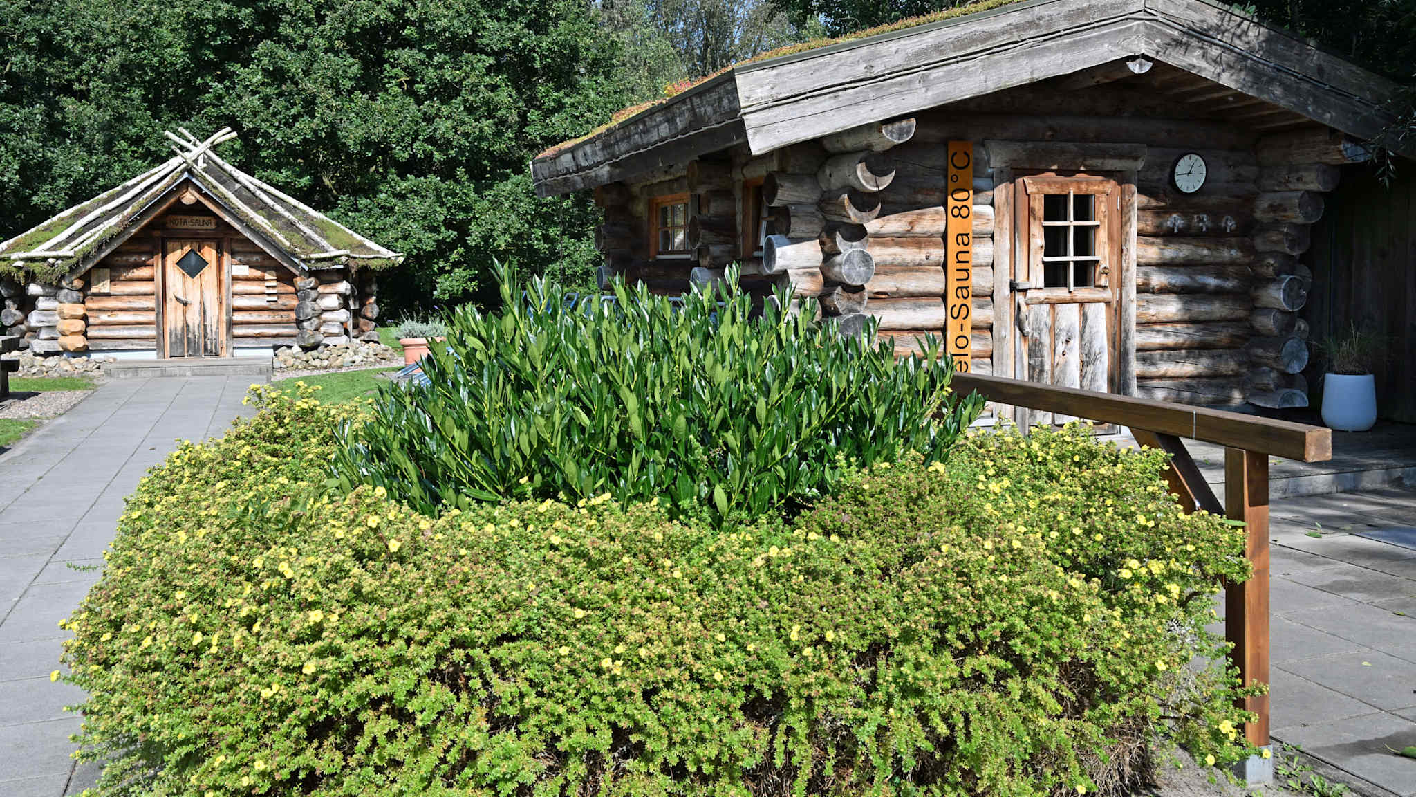 Zwei rustikale Saunahütten aus Rundholz im grünen Außenbereich des Nautimo Wilhelmshaven, eingerahmt von Pflanzen und Bäumen, mit Natursteinwegen und Holzdetails.