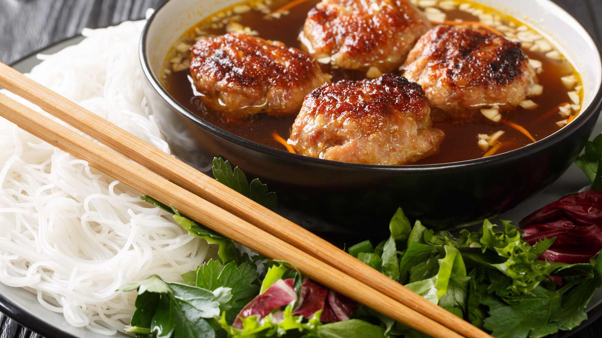 Ein Teller mit Salat, Kräutern und Reisnudeln und einer Schüssel mit vietnamesischen Bún chả in einer Brühe.