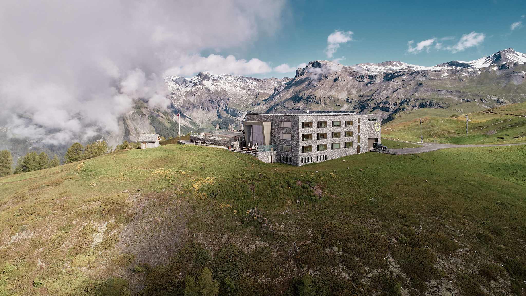 Designhotel Chetzeron im Wallis auf einer Bergwiese gelegen, umgeben von alpiner Landschaft mit Gipfeln und Wolken.
