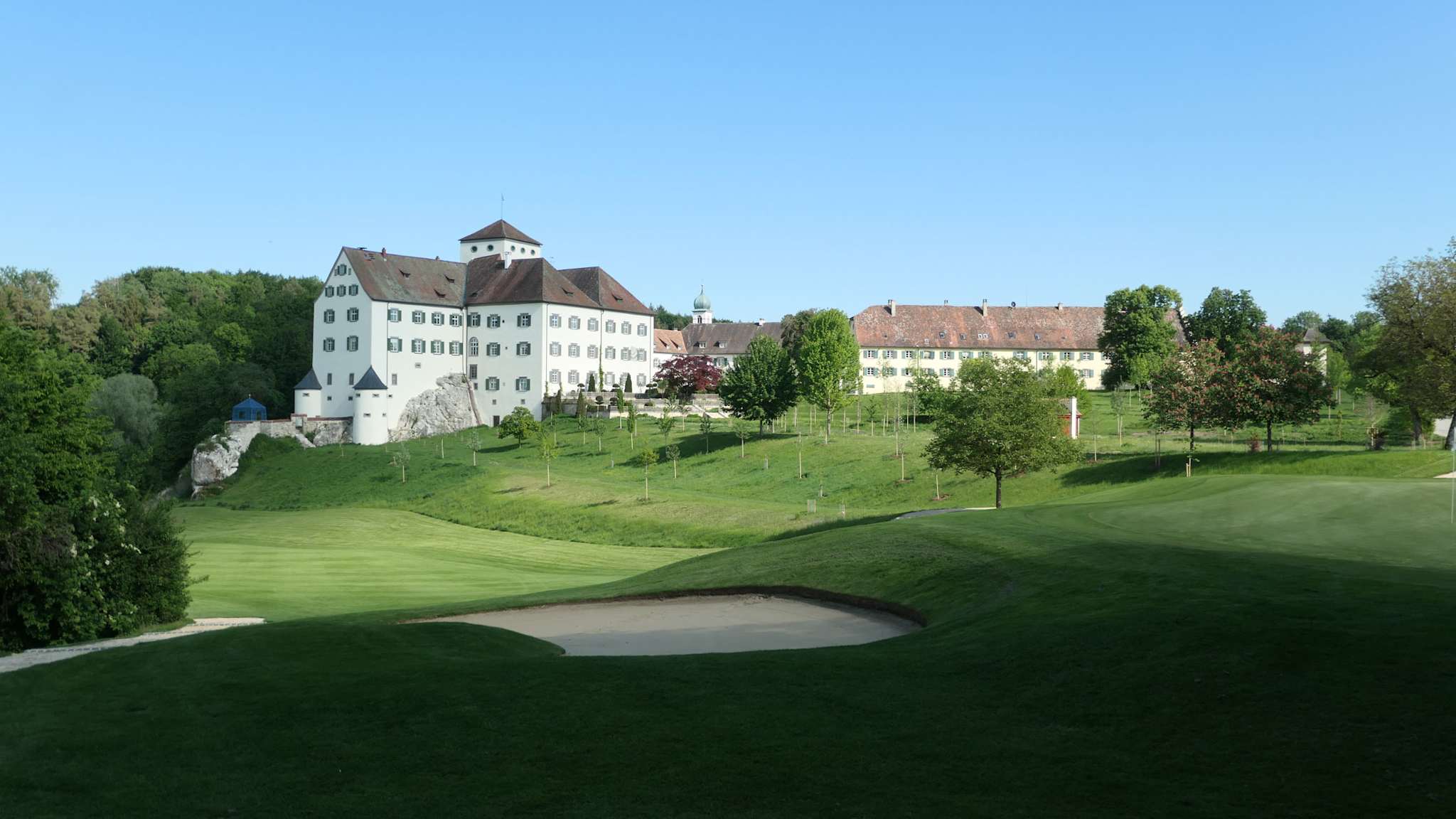 Hügelige Golfbahn mit Sandbunker im Vordergrund, im Hintergrund das helle Schloss Langenstein mit Renaissance-Elementen und umgebendem Grün