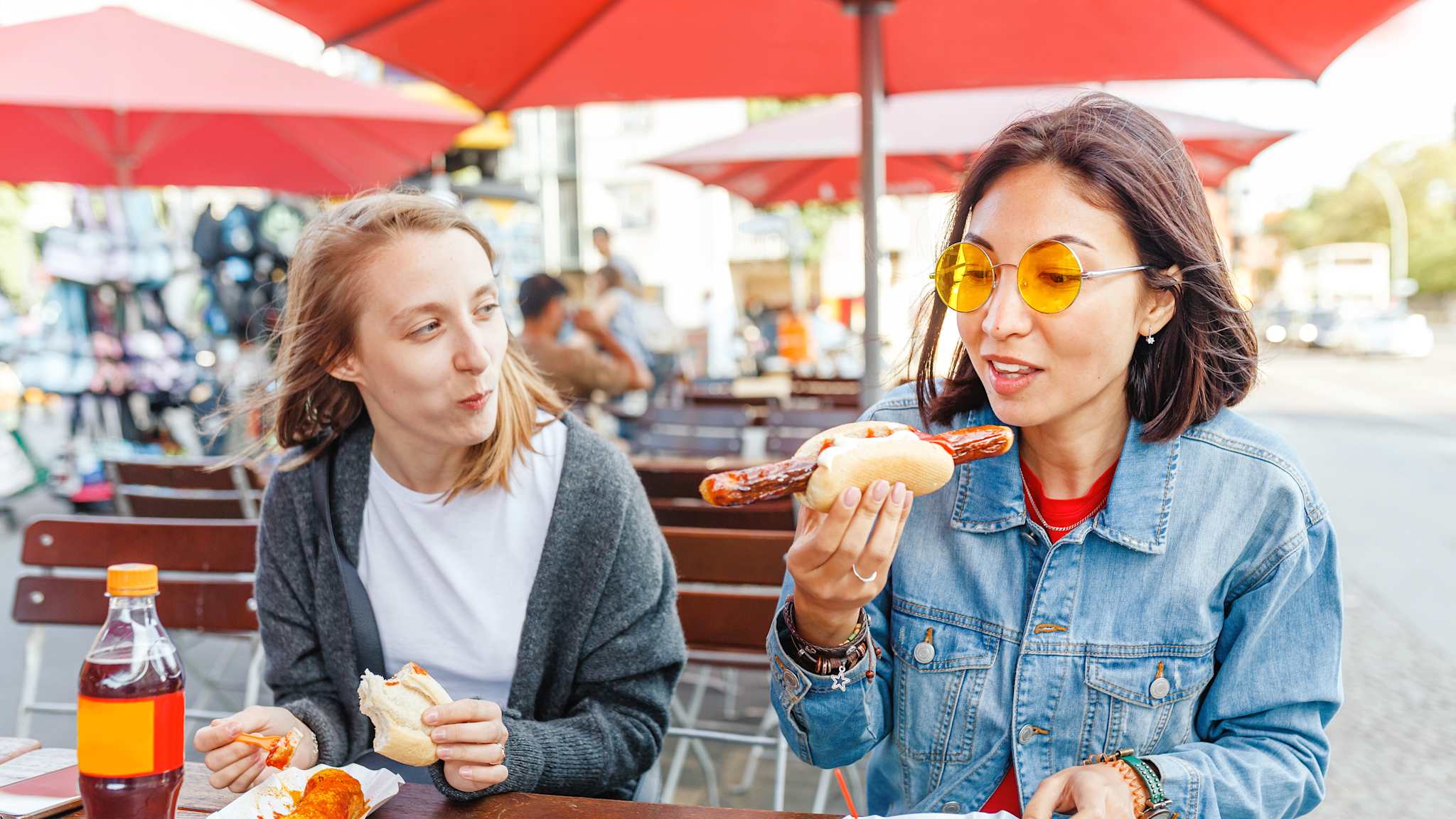 Zwei Frauen sitzen an einem Tisch unter roten Schirmen an einem Imbiss, die eine isst Currywurst mit Brötchen und die andere Bratwurst mit Pommes.