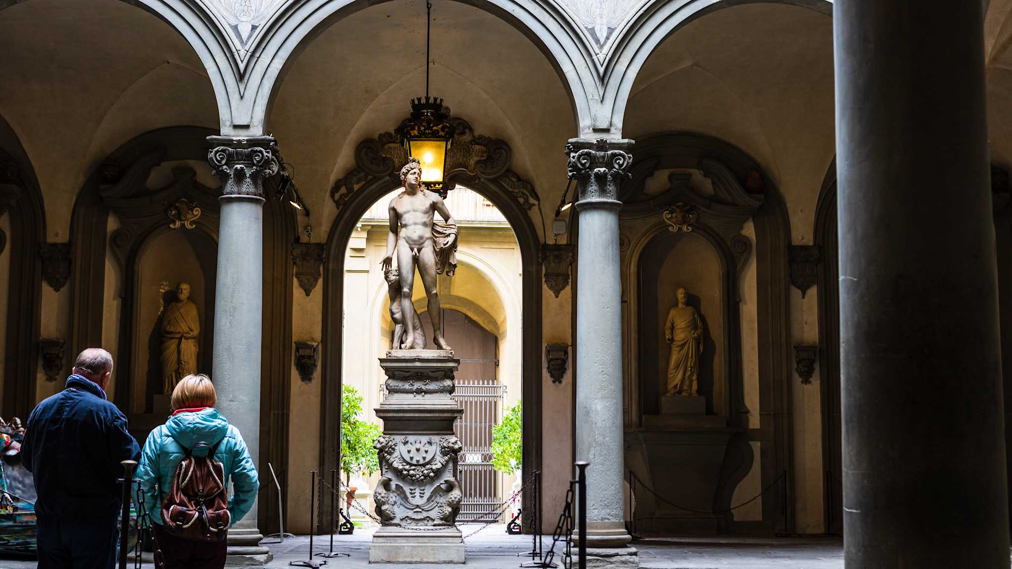Michelangelos David steht zwischen zwei Säulen in einem Bogengang in der Galleria dell'Accademia in Florenz. An der Rückwand hinter ihm stehen andere alte Statuen in einzelnen Wandnischen.