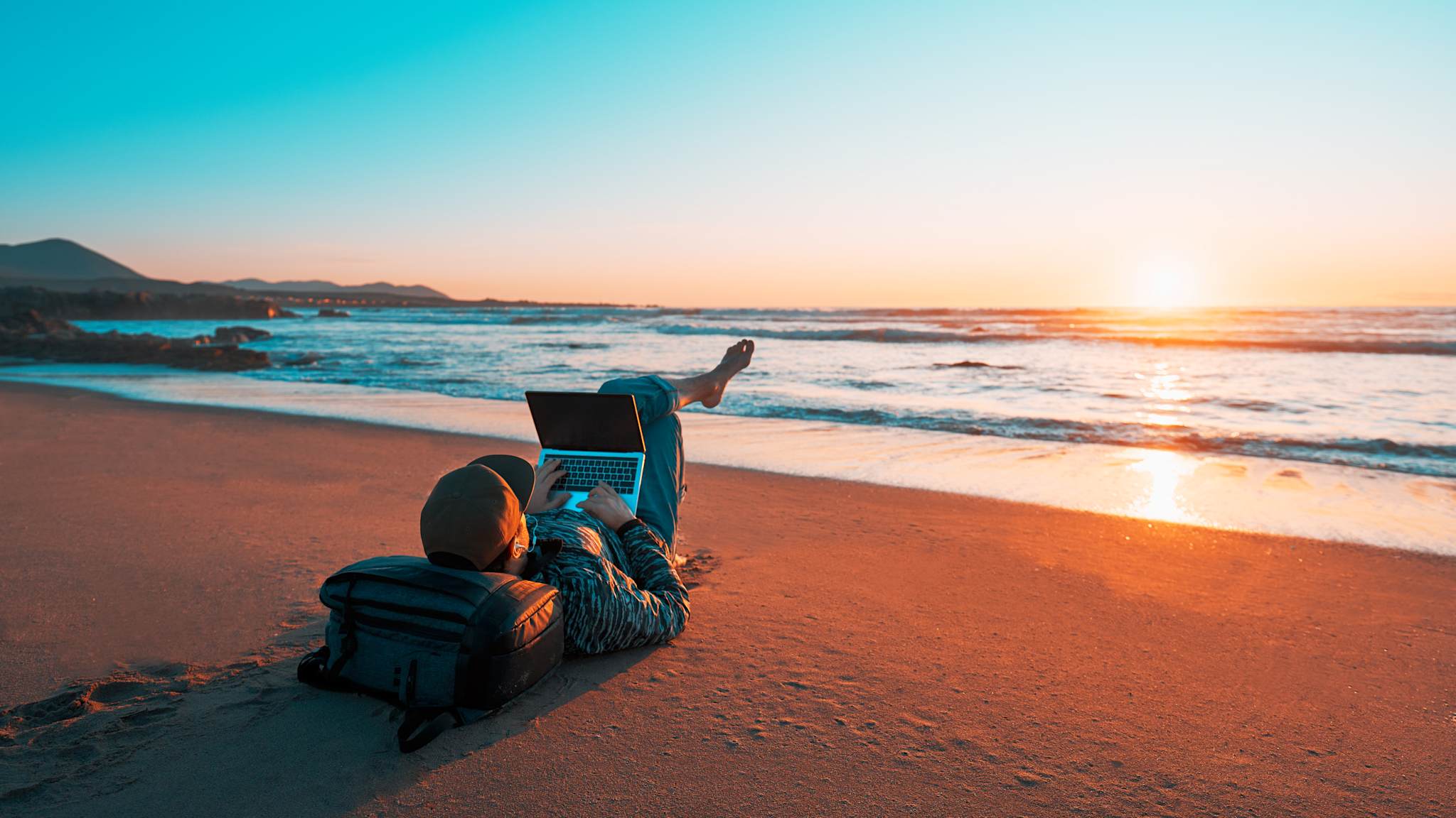 Mann liegt bei Sonnenuntergang am Strand, mit einem Laptop auf dem Schoß
