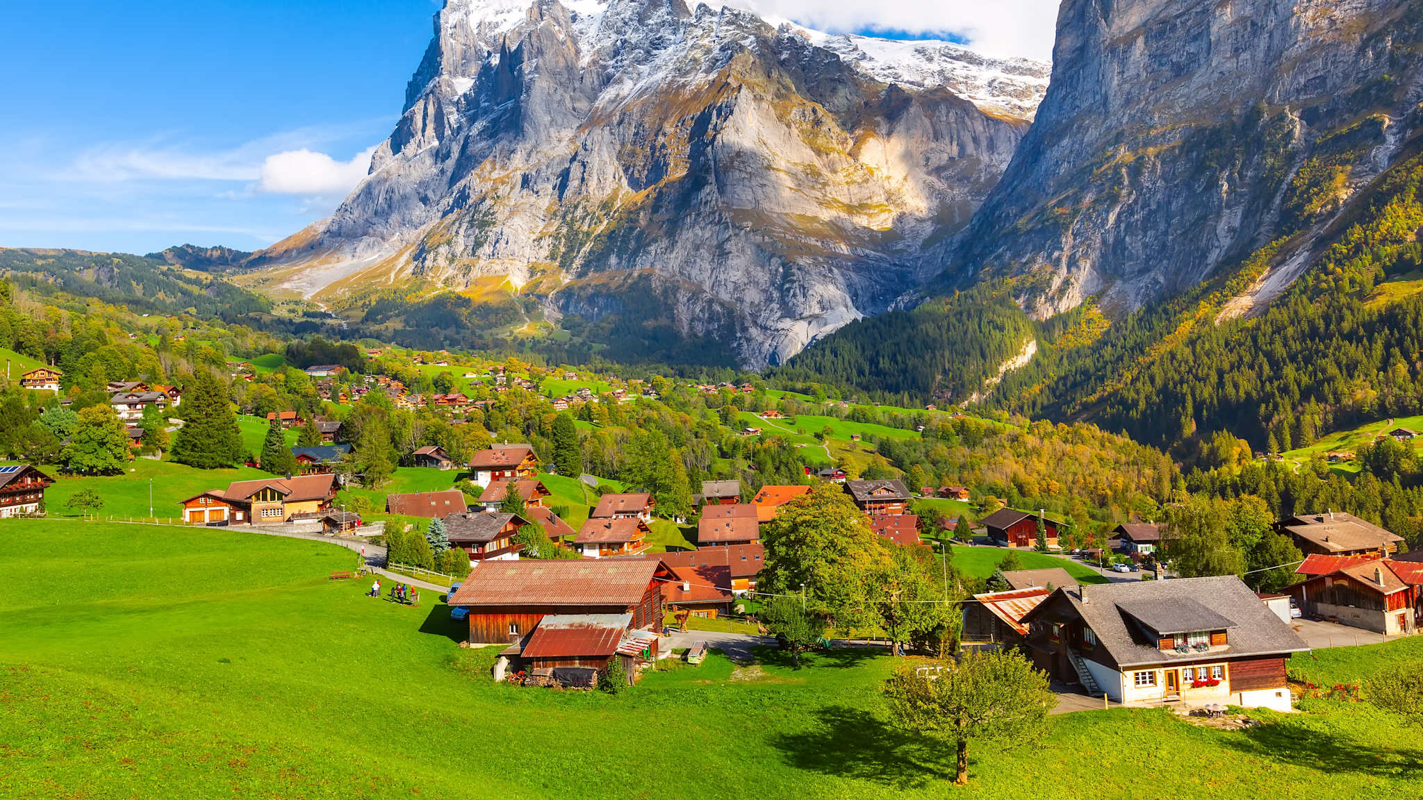 Ein kleines Dorf liegt in der sanften Hügellandschaft vor einem großen Gebirge.