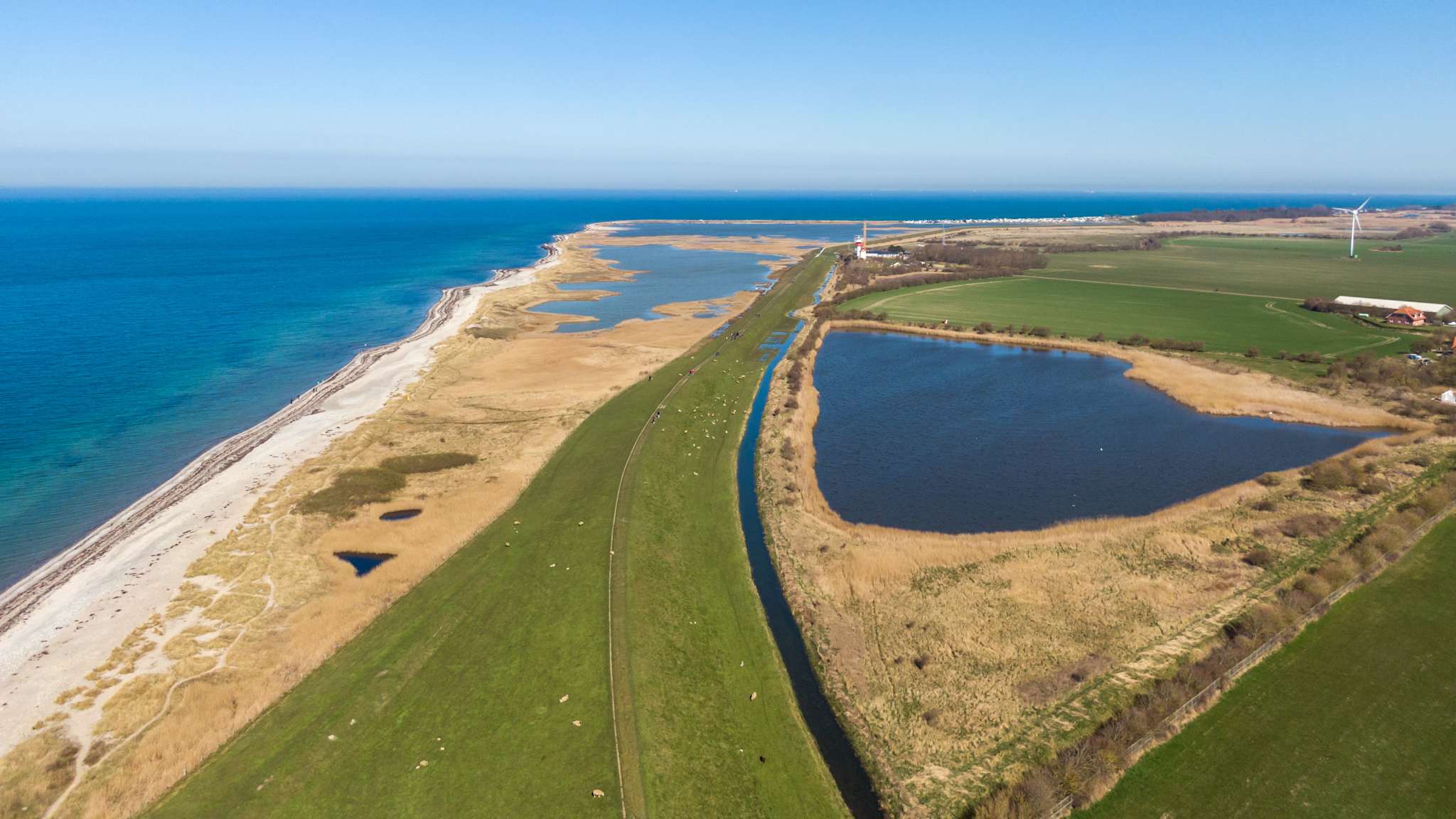Drohnenaufnahme über einen langen Strand und das Hinterland. Strand und Hinterland sind von großen Wasserflächen durchzogen.
