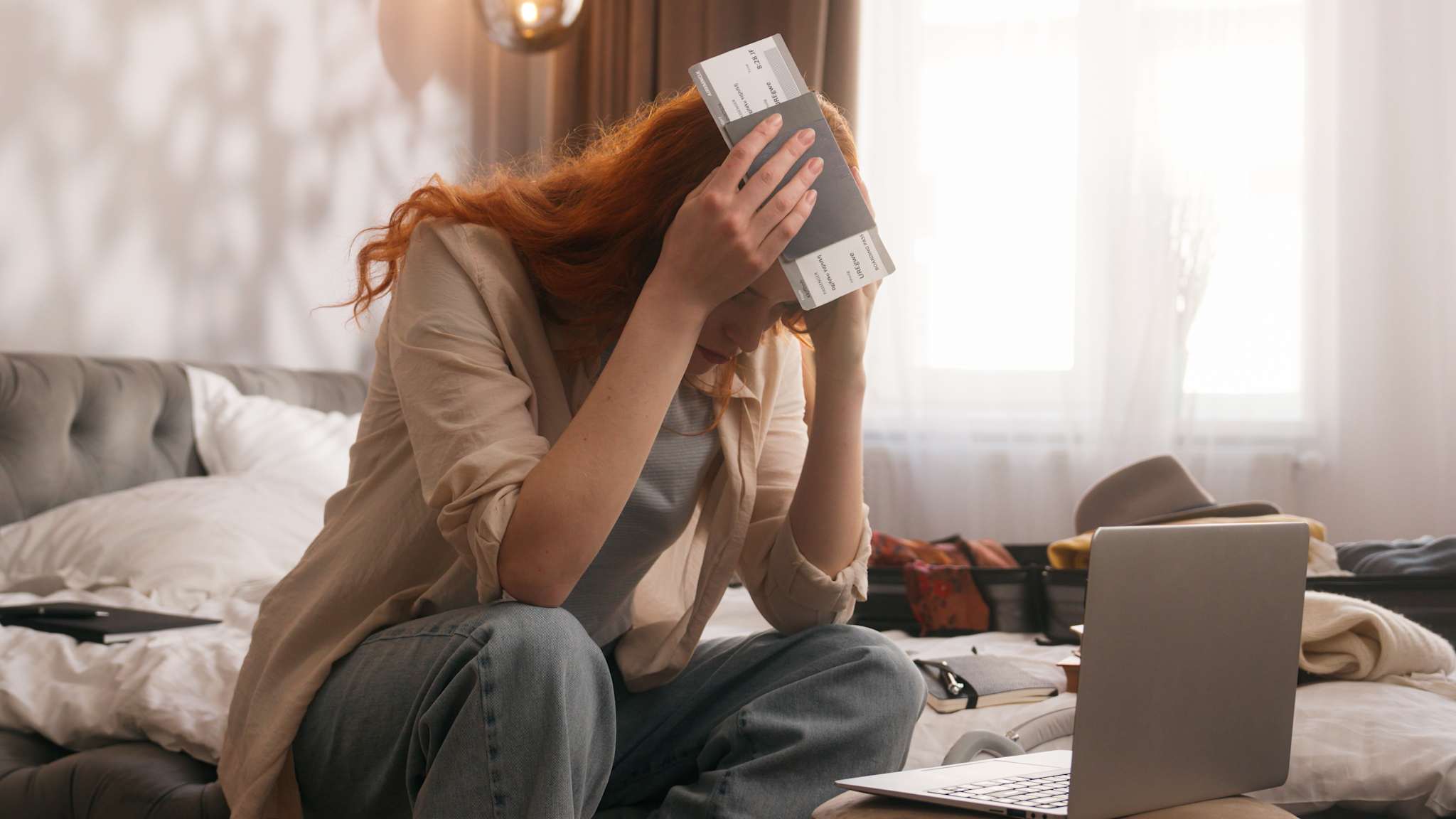 Frau mit Reisepass und Flugticket in der Hand hält sich den Kopf, dabei sitzt sie vor einem Laptop auf einem Bett