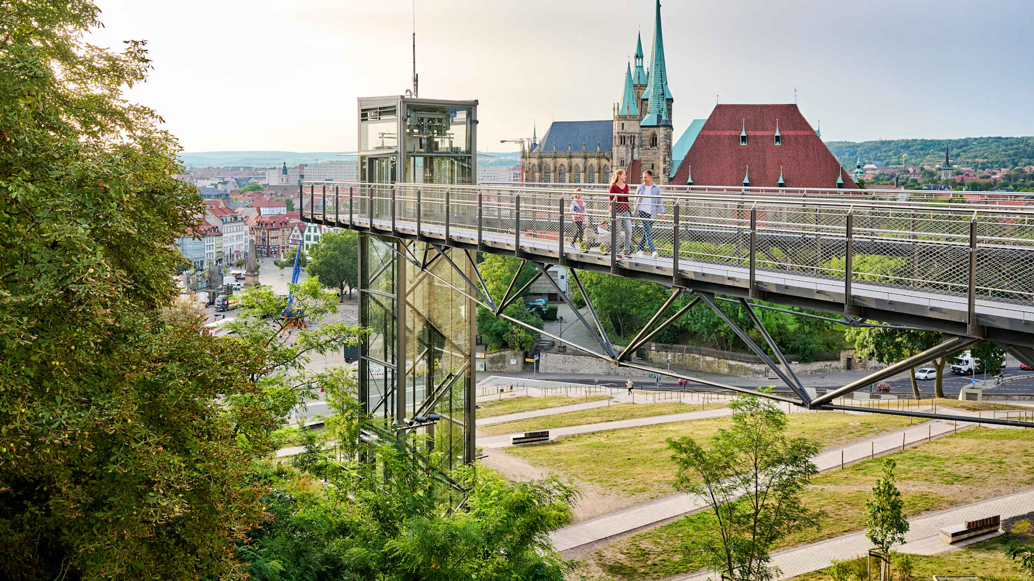 Ein Skywalk führt über einen Park, im Hintergrund liegt eine Kathedrale und eine Innenstadt mit historischen Fassaden