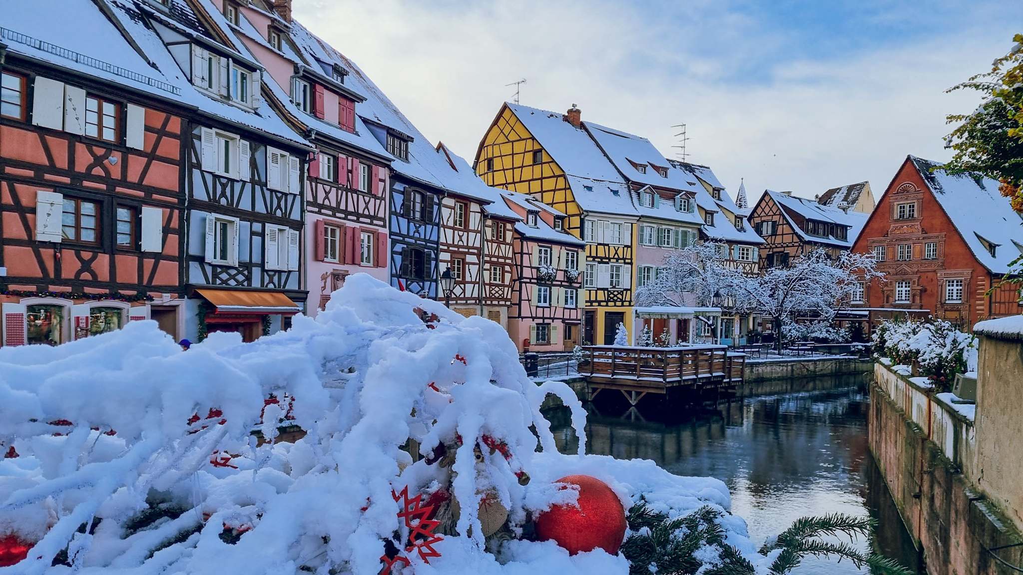 Winterliche Ansicht von Colmar mit bunt gestrichenen Fachwerkhäusern am Kanal, im Vordergrund verschneite Weihnachtsdekoration.