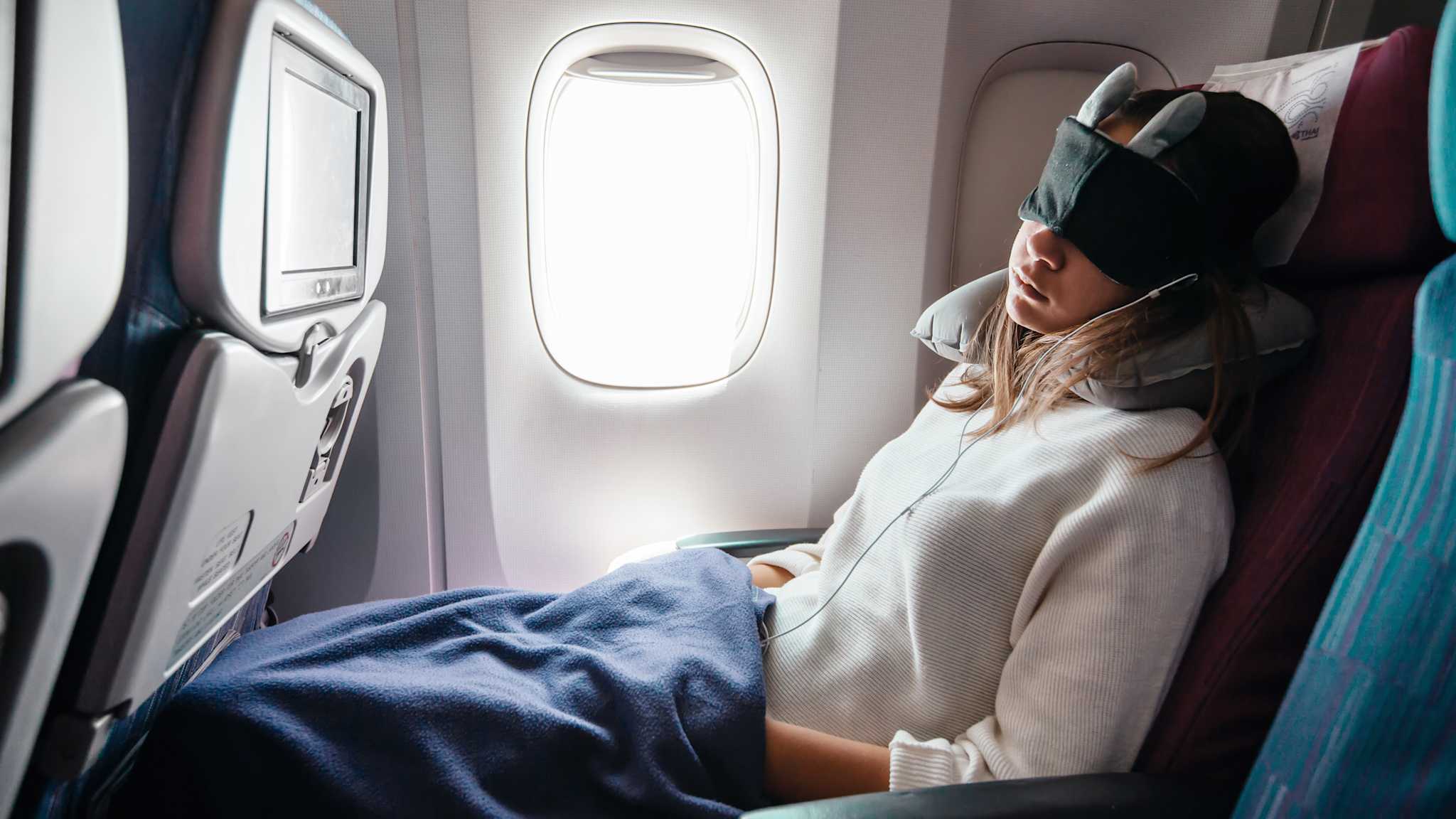 Frau schläft im Flugzeug mit Augenmaske, Nackenkissen und Decke