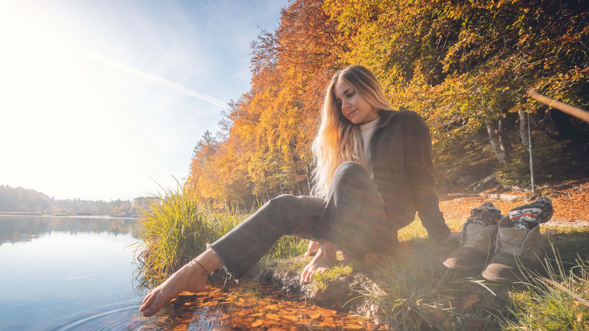 Frau sitzt am herbstlichen Seeufer und taucht die Füße ins Wasser, während bunte Wälder und sanftes Licht eine stille Atmosphäre schaffen.