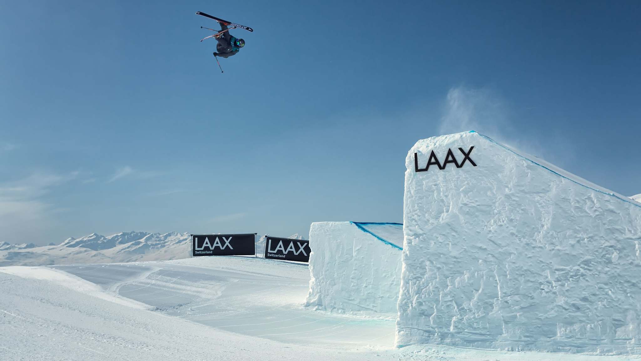 Freestyle-Skifahrer in der Luft bei einem Trick über einen Big-Air-Kicker im Snowpark Laax, blauer Himmel und verschneite Alpen im Hintergrund.