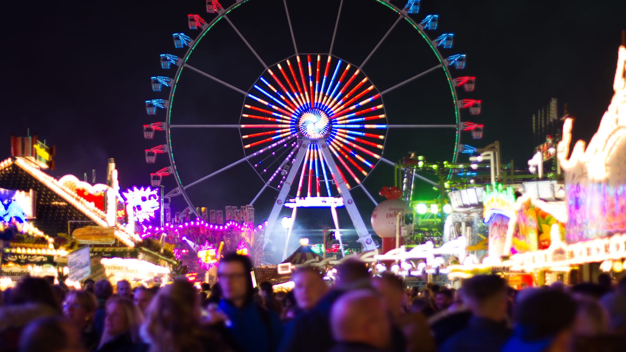 Ein Riesenrad auf einem Volksfest leuchtet bei Nacht. Davor stehen viele Menschen zwischen Buden.