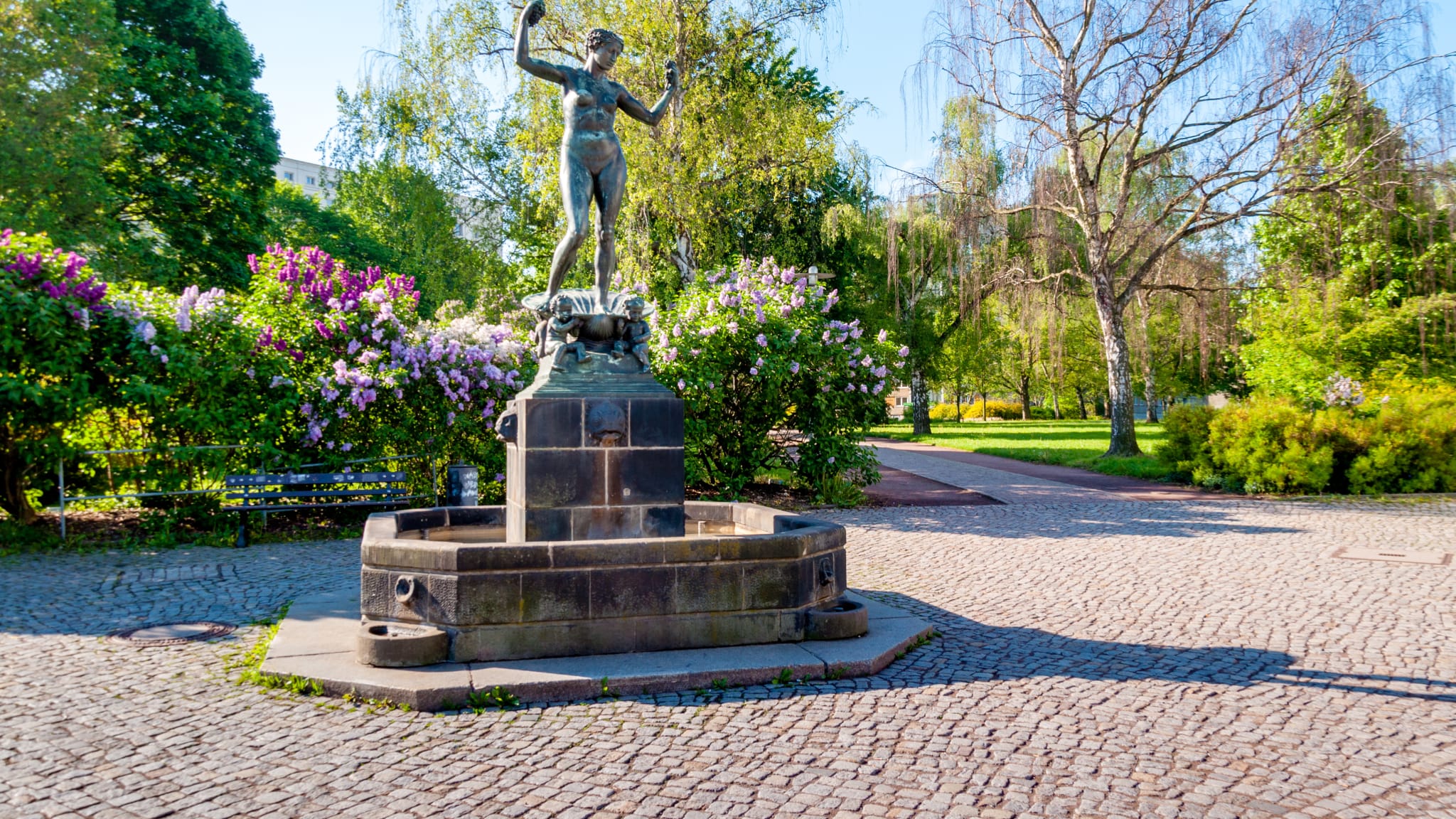 Man sieht einen Brunnen mit der Statue einer unbekleideten Frau. Dahinter stehen einige Bäume und eine Hecke mit bunt blühenden Büschen.
