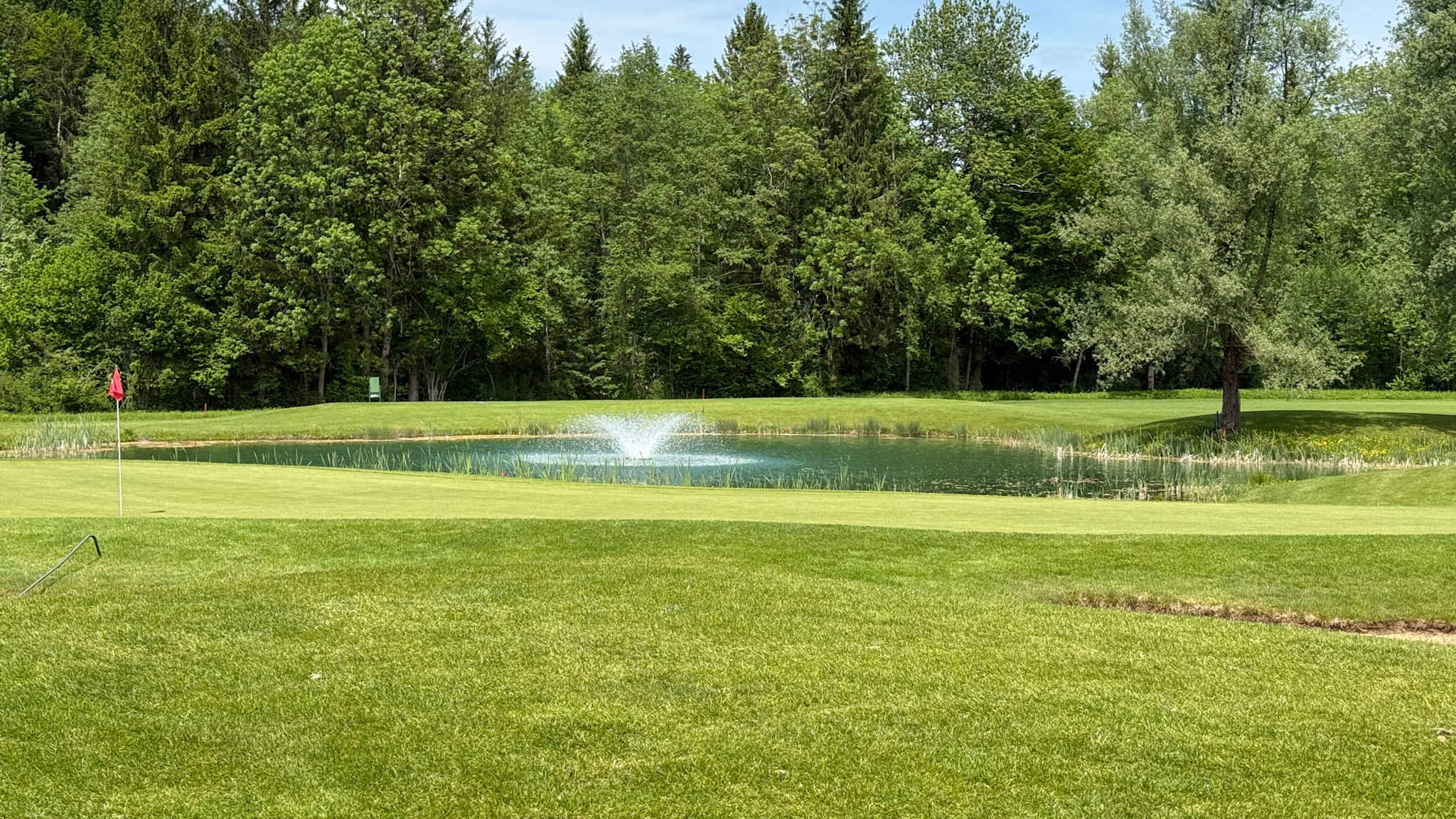 Grünes Putting Green mit roter Fahne, davor ein kleiner Teich mit Wasserfontäne, im Hintergrund dichter Mischwald im Golfpark Bregenzerwald in Österreich