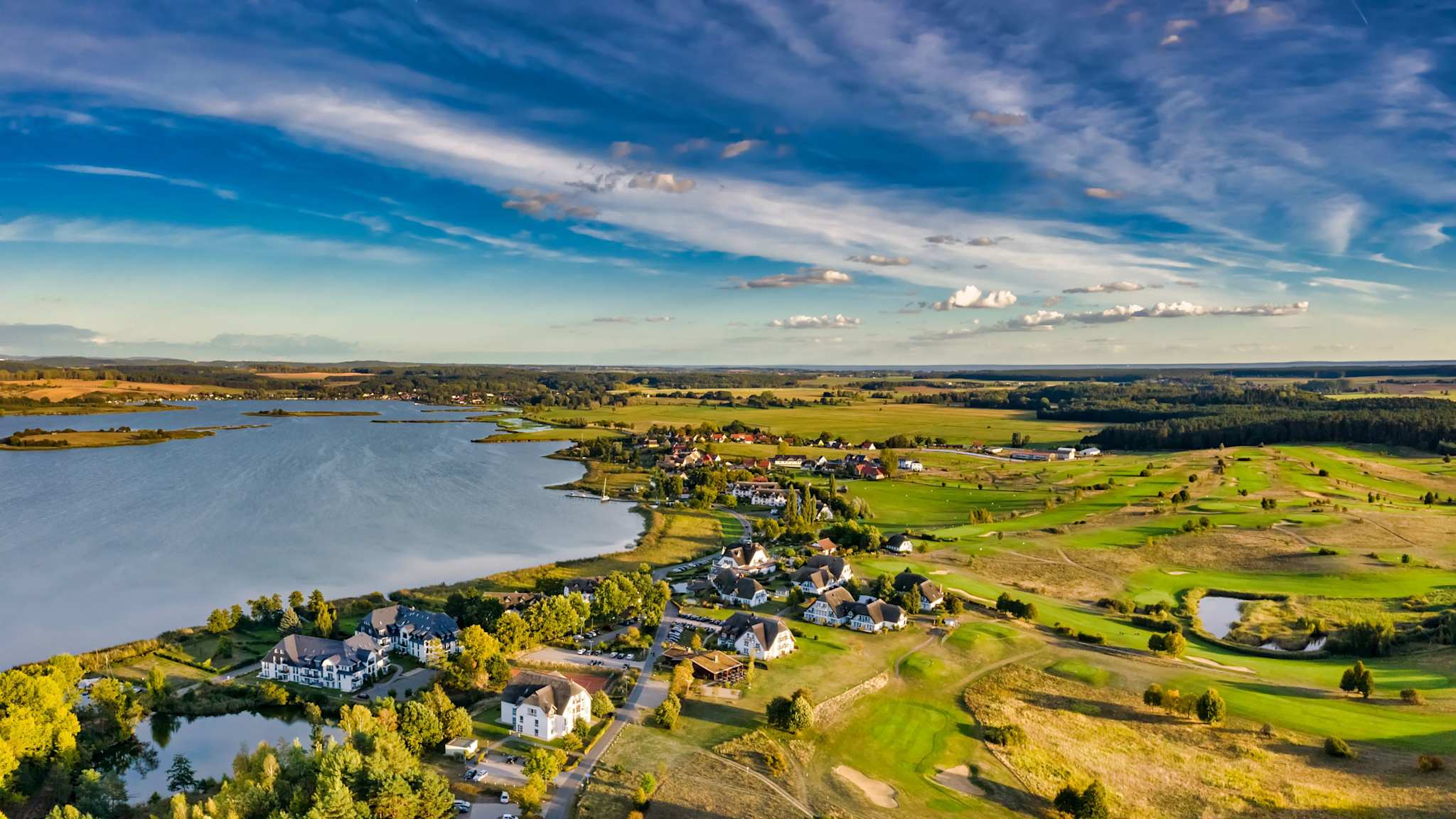 Luftaufnahme des Golfparks Balmer See mit Blick auf das Achterwasser, gepflegten Fairways, Reetdachhäusern und weiter Küstenlandschaft unter einem weiten Himmel