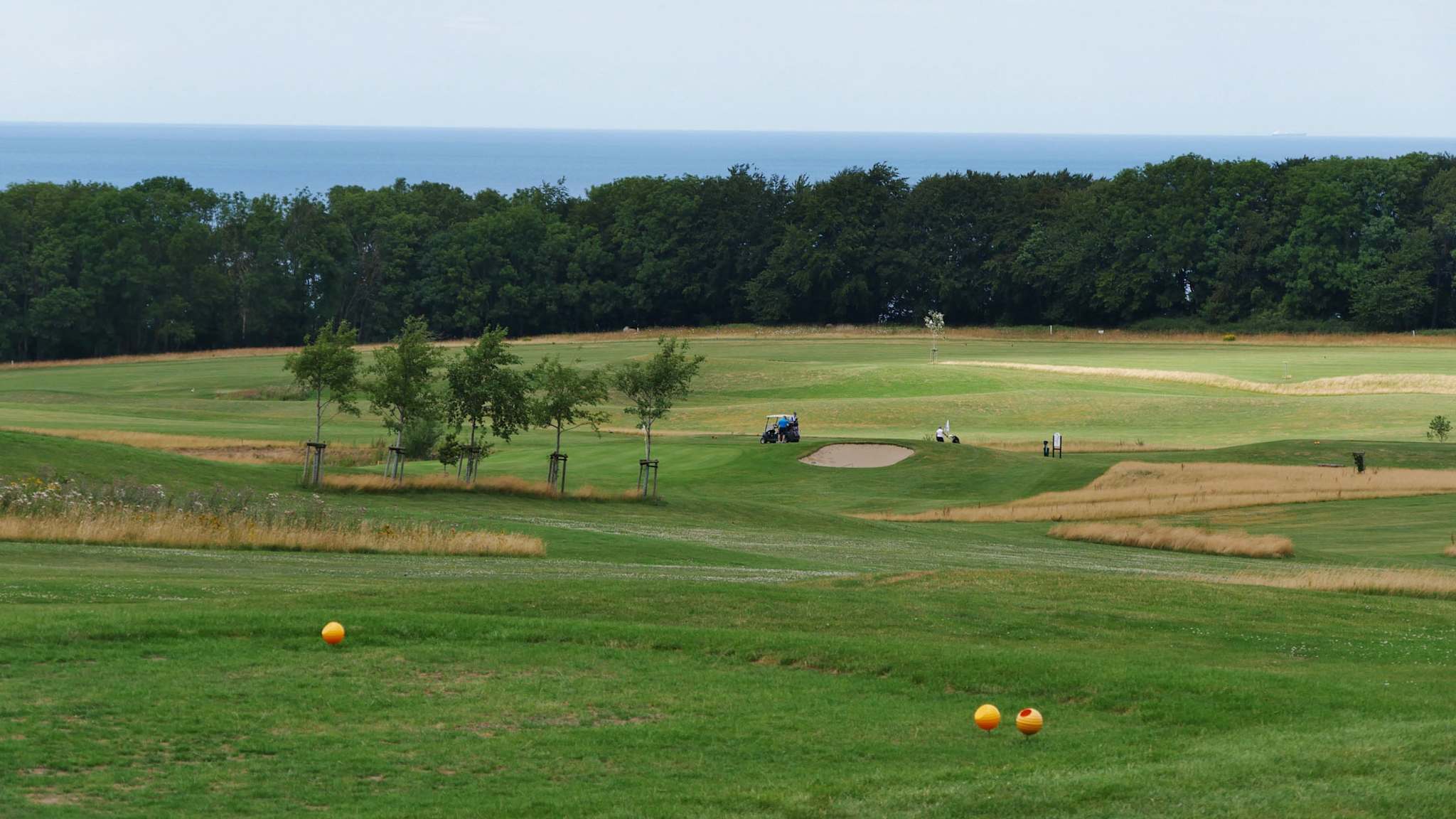 Weitläufige Fairways des Golfplatzes Schloss Ranzow mit Blick auf die Ostsee, eingerahmt von Bäumen und sanften Hügeln, mit Golfcart und Bunker im Hintergrund
