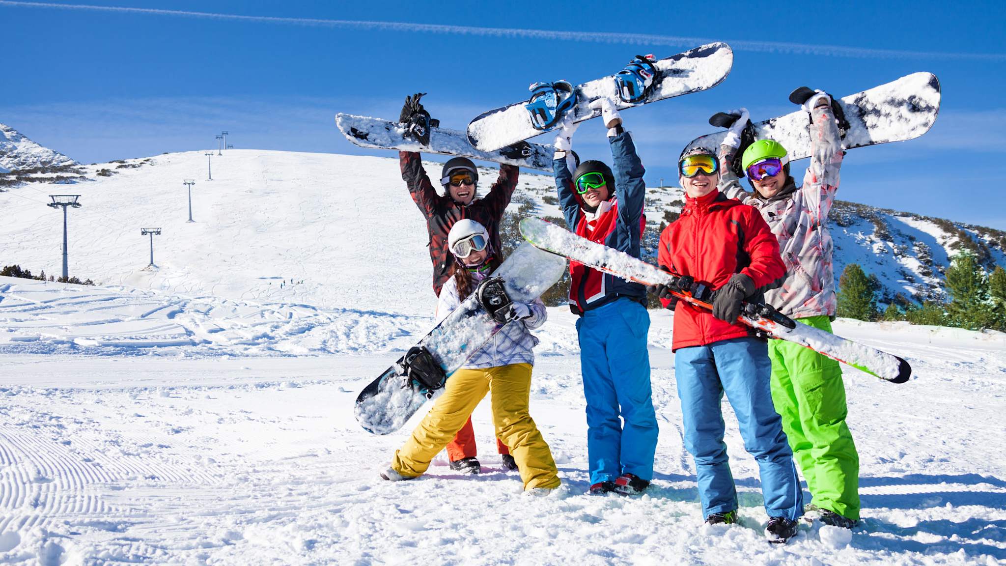 Gruppe junger Menschen mit Snowboards jubelt im Sonnenschein auf einer Skipiste.