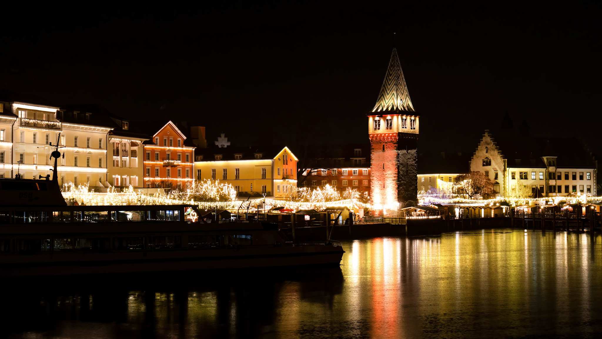 Nächtliche Ansicht der Lindauer Hafenweihnacht mit beleuchteten Häusern, Marktständen und dem angestrahlten Mangturm; die Lichter spiegeln sich deutlich im ruhigen Wasser des Bodensees.
