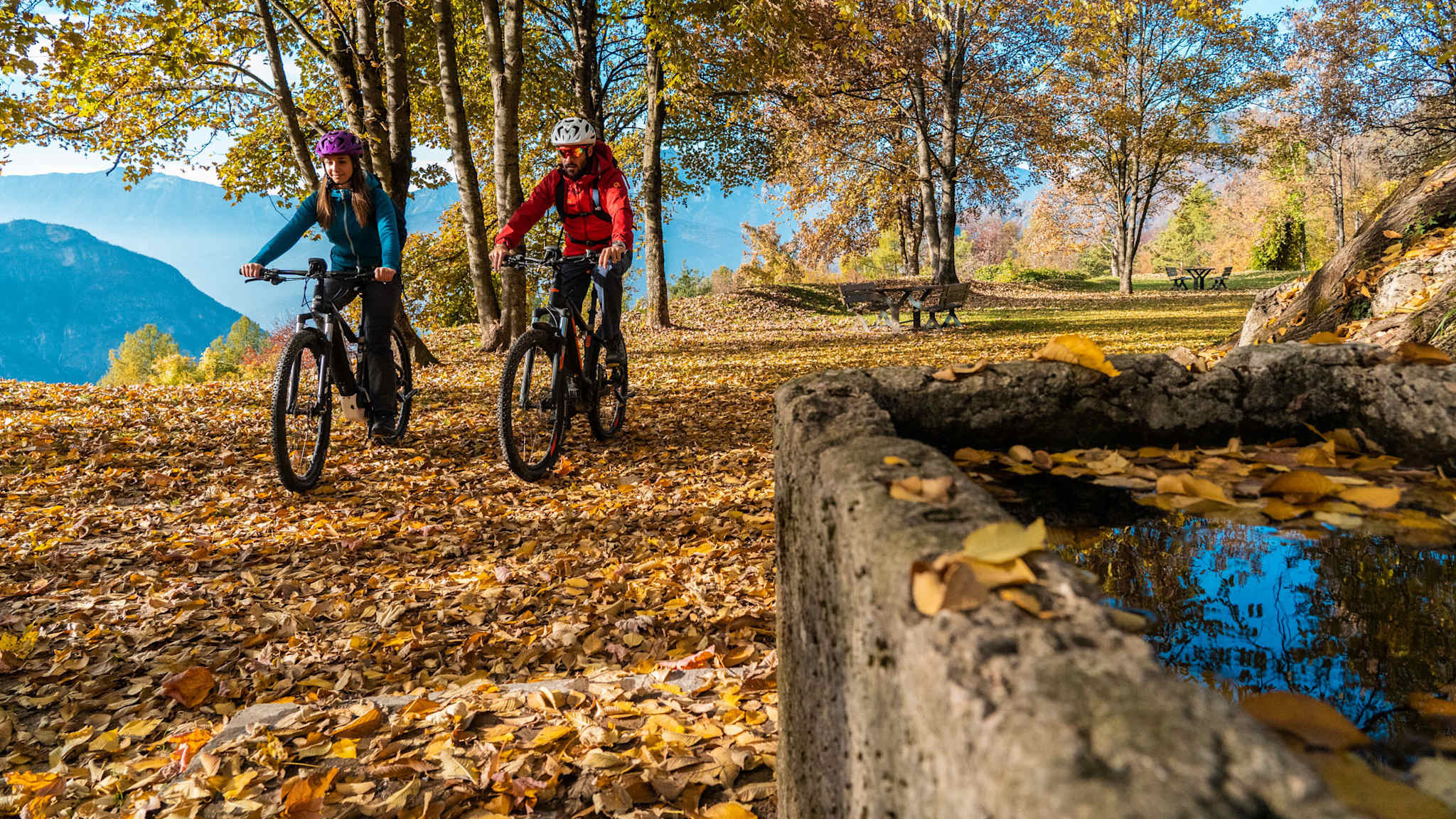 Zwei Radfahrer fahren durch einen mit buntem Herbstlaub bedeckten Waldweg, daneben ein alter steinerner Trog mit Wasser; im Hintergrund Bäume und weite Berglandschaft.