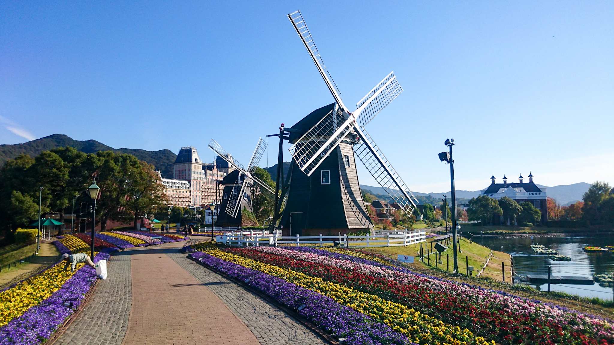 Von bunten Blumen gesäumter Weg führt an großen holländischen Mühlen und Grachten vorbei auf größere Gebäude zu im Freizeitpark Huis Ten Bosch in Nagasaki, Japan.