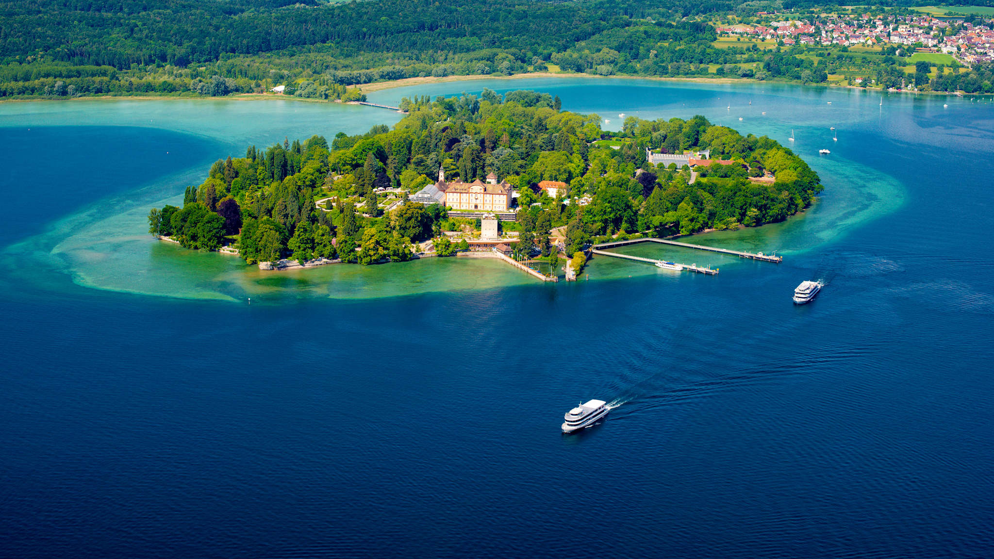 Blick auf eine kleine, bewaldete Insel von oben, drumherum blaues Meer, im Hintergrund das Festland