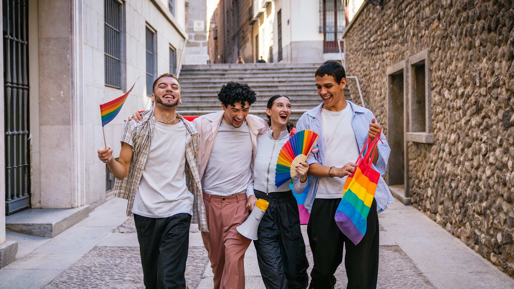 Vier junge Menschen gehen durch eine Straße und halten eine Fahne, einen Fächer und eine Tasche in den Pride-Farben.