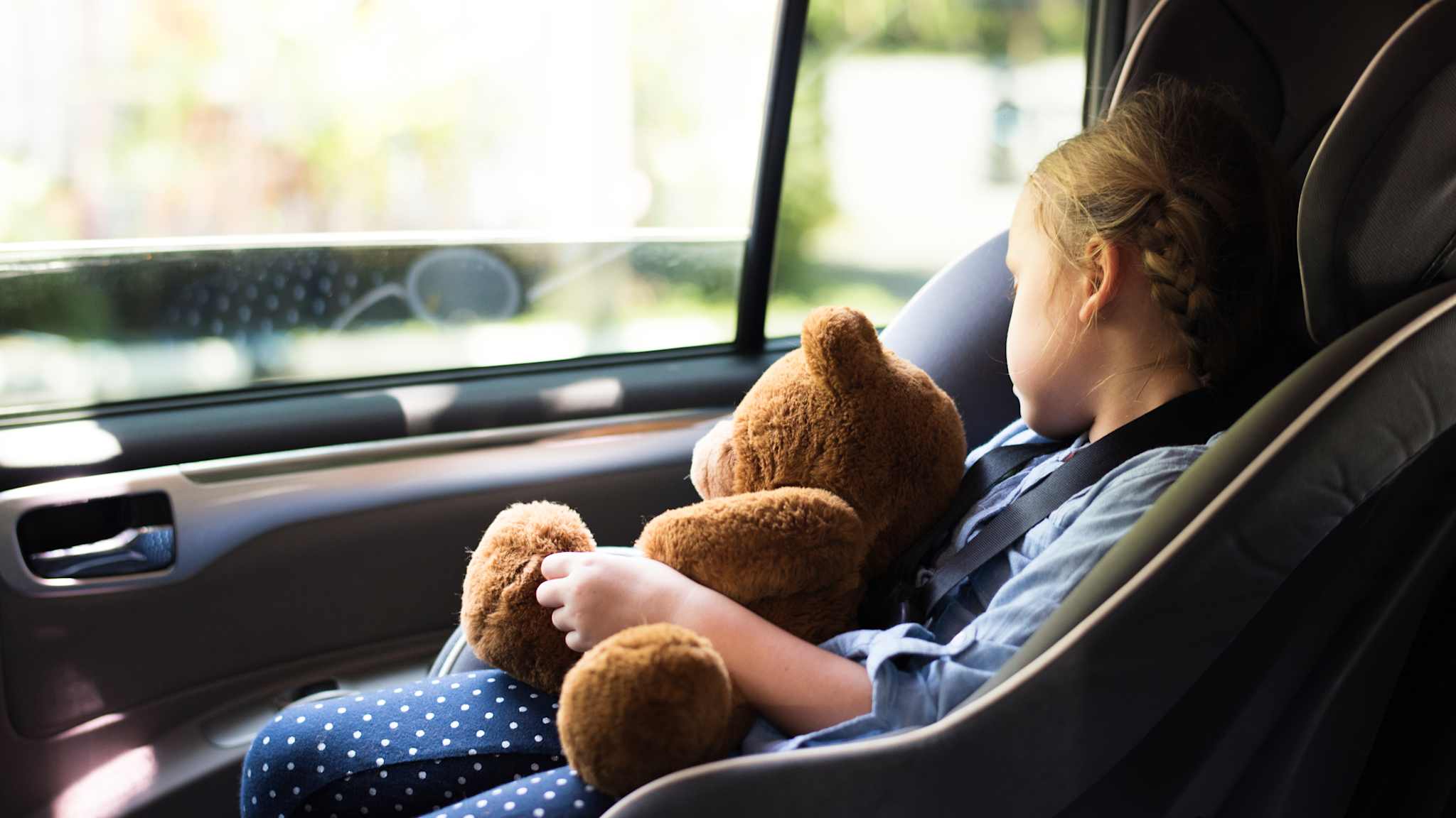 Ein Kind schläft im Kindersitz auf dem Rücksitz am Fenster mit einem Teddy im Arm.