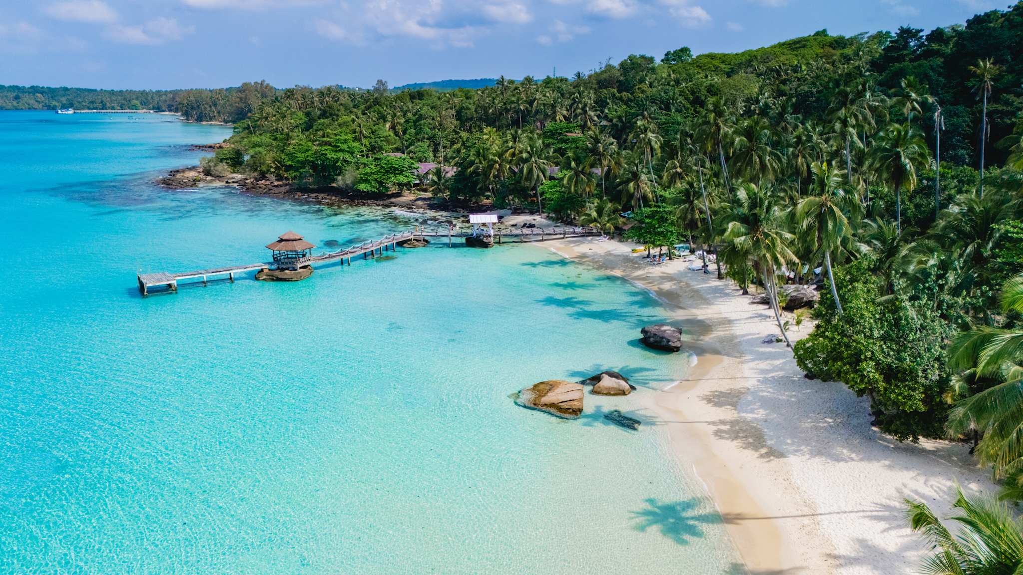 Strand der thailändischen Insel Koh Kood mit helltürkisenem Wasser, einem kleinen, einfachen Holzsteg und einem Palmenwald.