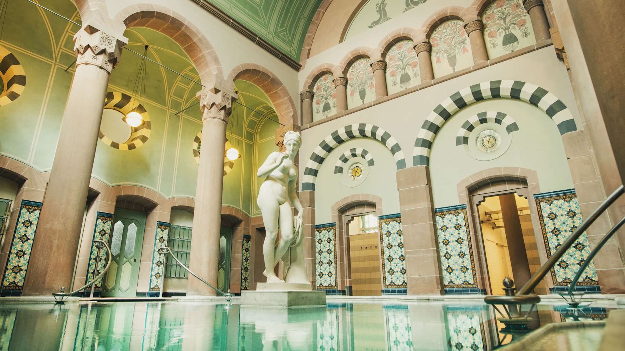 Eleganter Thermalraum mit großer Marmorstatue in einem Becken, umgeben von filigran bemalten Rundbögen, hohen Säulen, Jugendstil-Ornamentik und farbigen Wandfliesen im Palais Thermal Bad Wildbad.