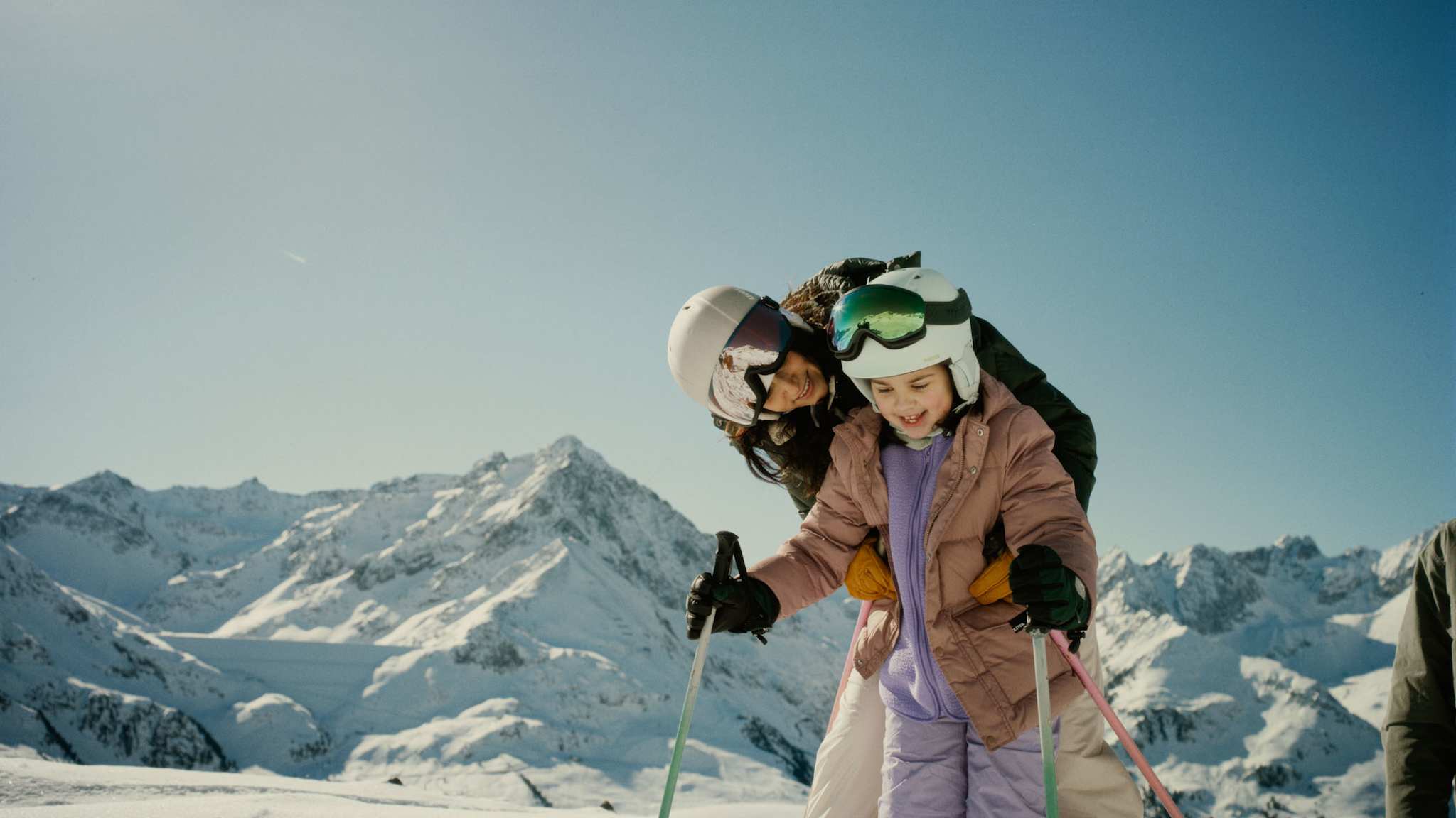 Eine Mutter hilft ihrem Kind beim Skifahren in Kühtai, beide lachen, im Hintergrund die verschneiten Gipfel der Tiroler Alpen.