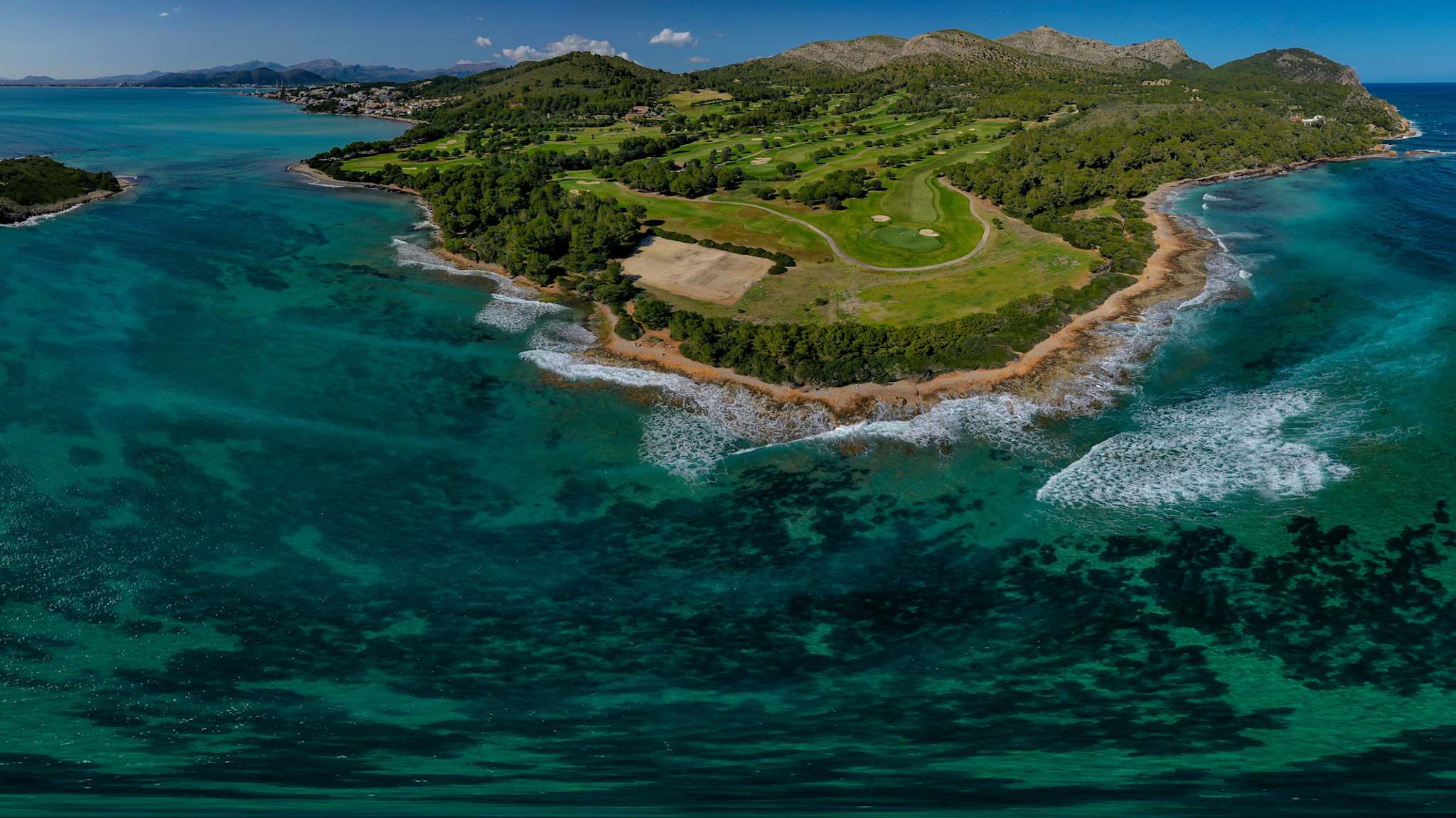 Luftaufnahme des Club de Golf Alcanada auf einer Halbinsel mit grünem Fairway, umgeben von türkisfarbenem Meer und Wellen, mit Leuchtturminsel und Bergen im Hintergrund bei Alcúdia auf Mallorca.