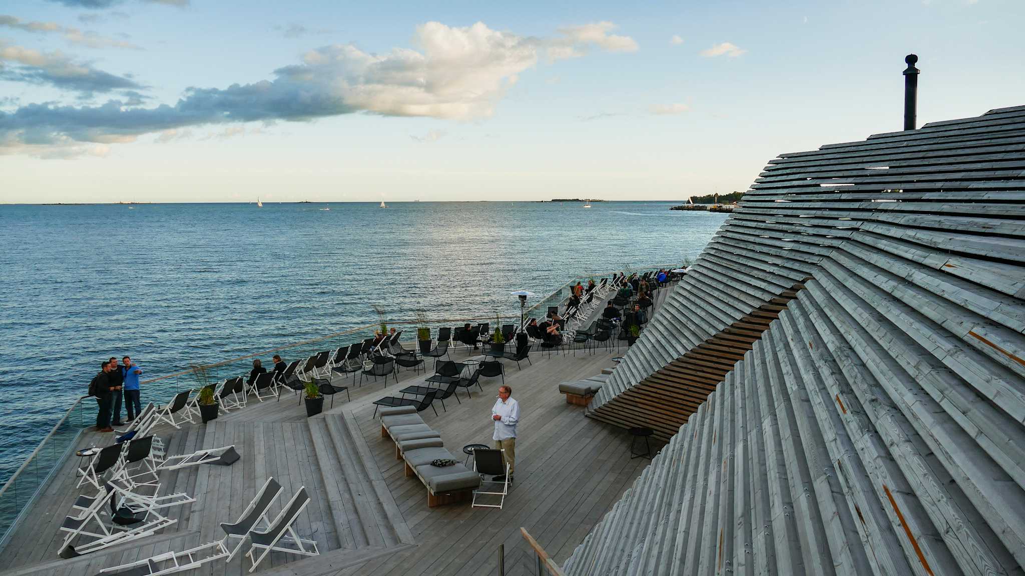 Menschen stehen auf den Holzterrassen eines großen Gebäudes am Meer.