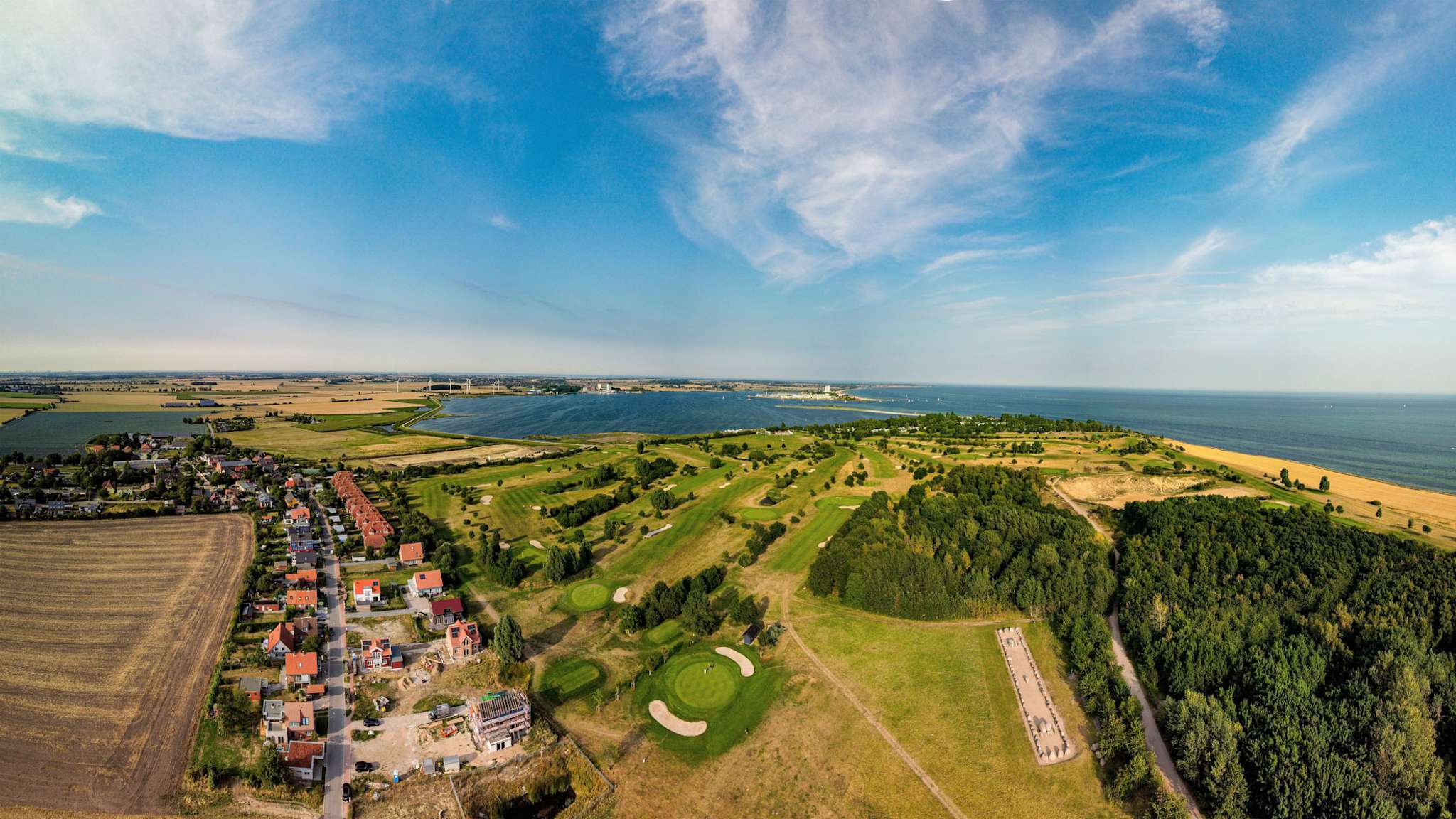 Weitwinkel-Luftbild des Golfparks Fehmarn mit direkter Lage an der Ostsee, grünen Fairways, Waldflächen, dem Ortsteil Wulfen und Blick auf den Burger Binnensee