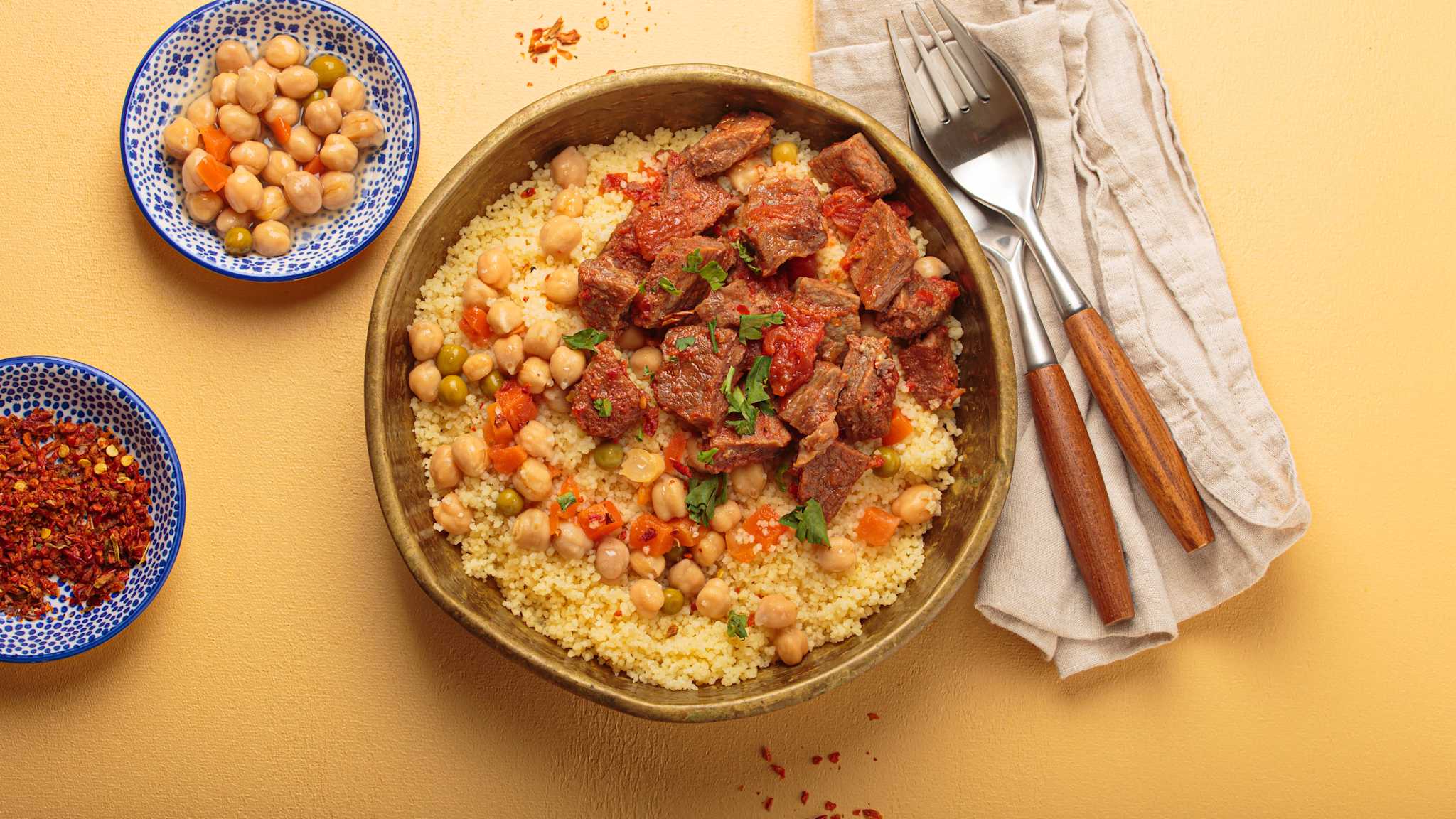 Eine Große Schale mit Couscous, links daneben zwei kleine Schalen mit Kichererbsen und Gewürz, rechts daneben Besteck und Serviette
