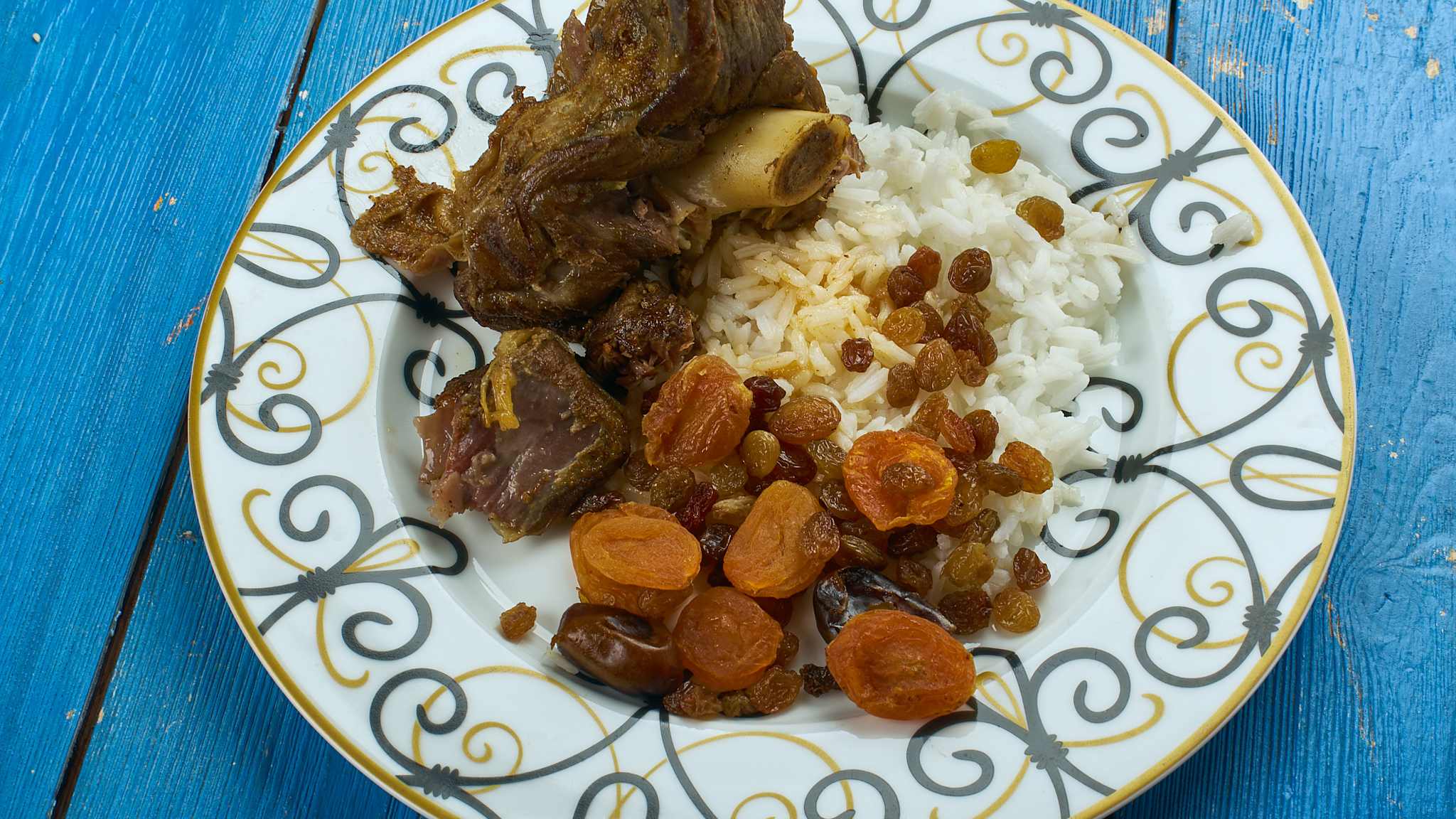 Teller auf blauem Holztisch, auf dem Teller liegen Reis, getrocknete Früchte und Lamm