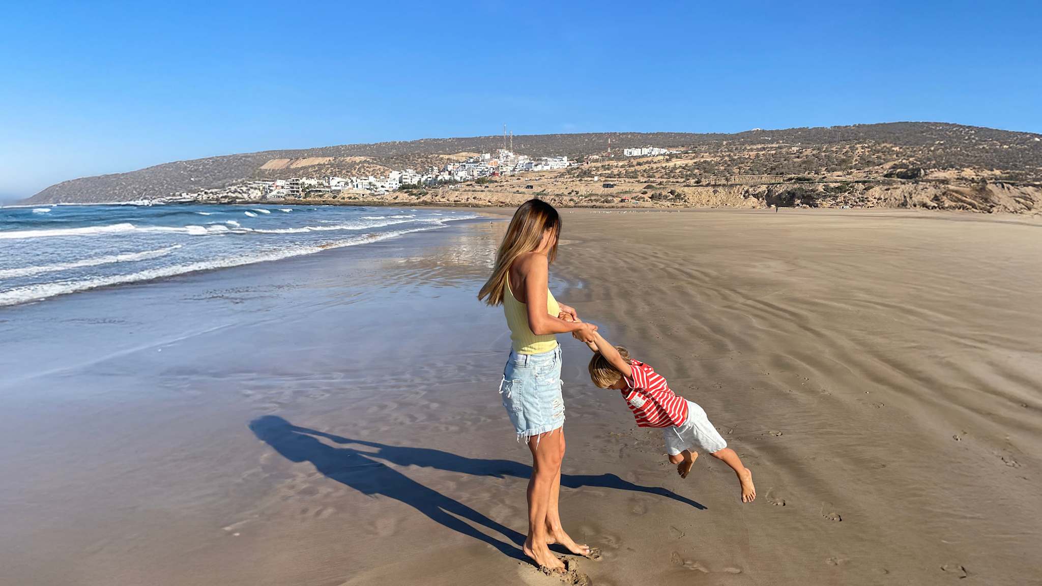 Eine Mutter spielt mit ihrem Sohn am Strand. Weit im Hintergrund erheben sich Hügel und ein Dorf.