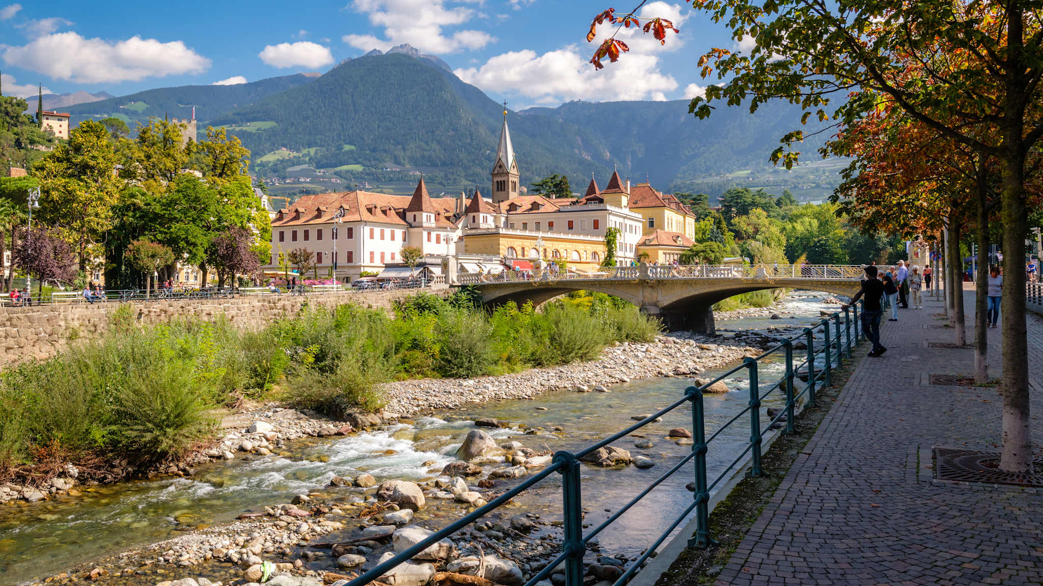 Spazierweg mit kleiner Allee am Fluss Passer mit Ausblick auf die Stadt Meran und die Berge im Hintergrund.