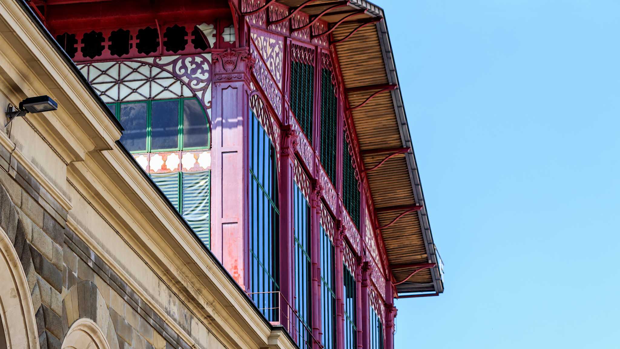Blick von unten zum Gebäude des Mercato Centrale in Florenz: Ein mit dunkelroten Panelen, verschnörkelter Ornamentik und grünen Fensterrahmen verkleideter Aufbau mit Dachgiebel auf ein klassisches Steingebäude mit runden Durchgängen.
