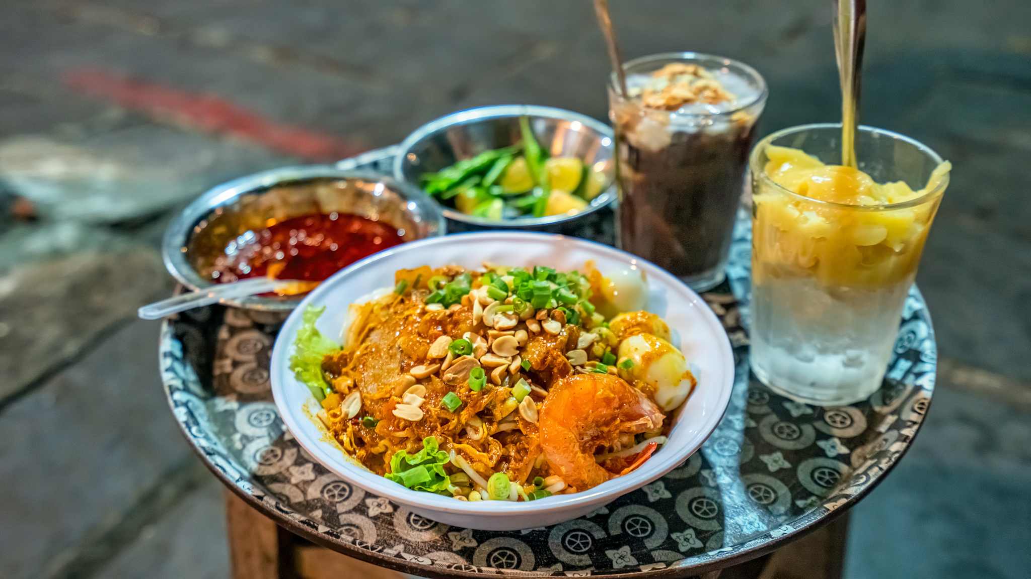 Auf einem kleinen Beistelltisch steht ein Teller vietnamesisches Mi Quảng, eine Soße und Getränke.