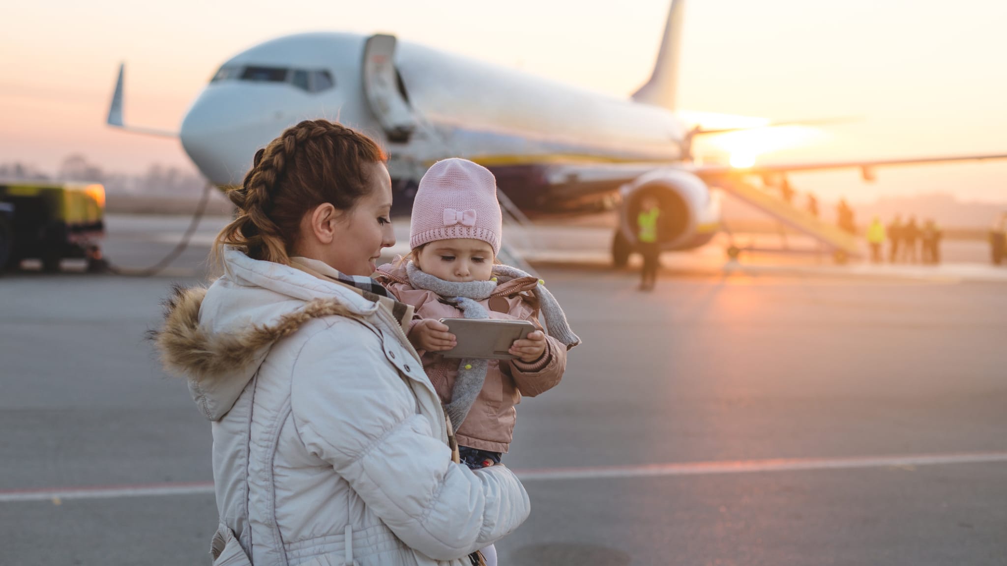 Mutter steht vor einem Flugzeug mit ihrer kleinen Tochter auf dem Arm © iStock.com/FluxFactory